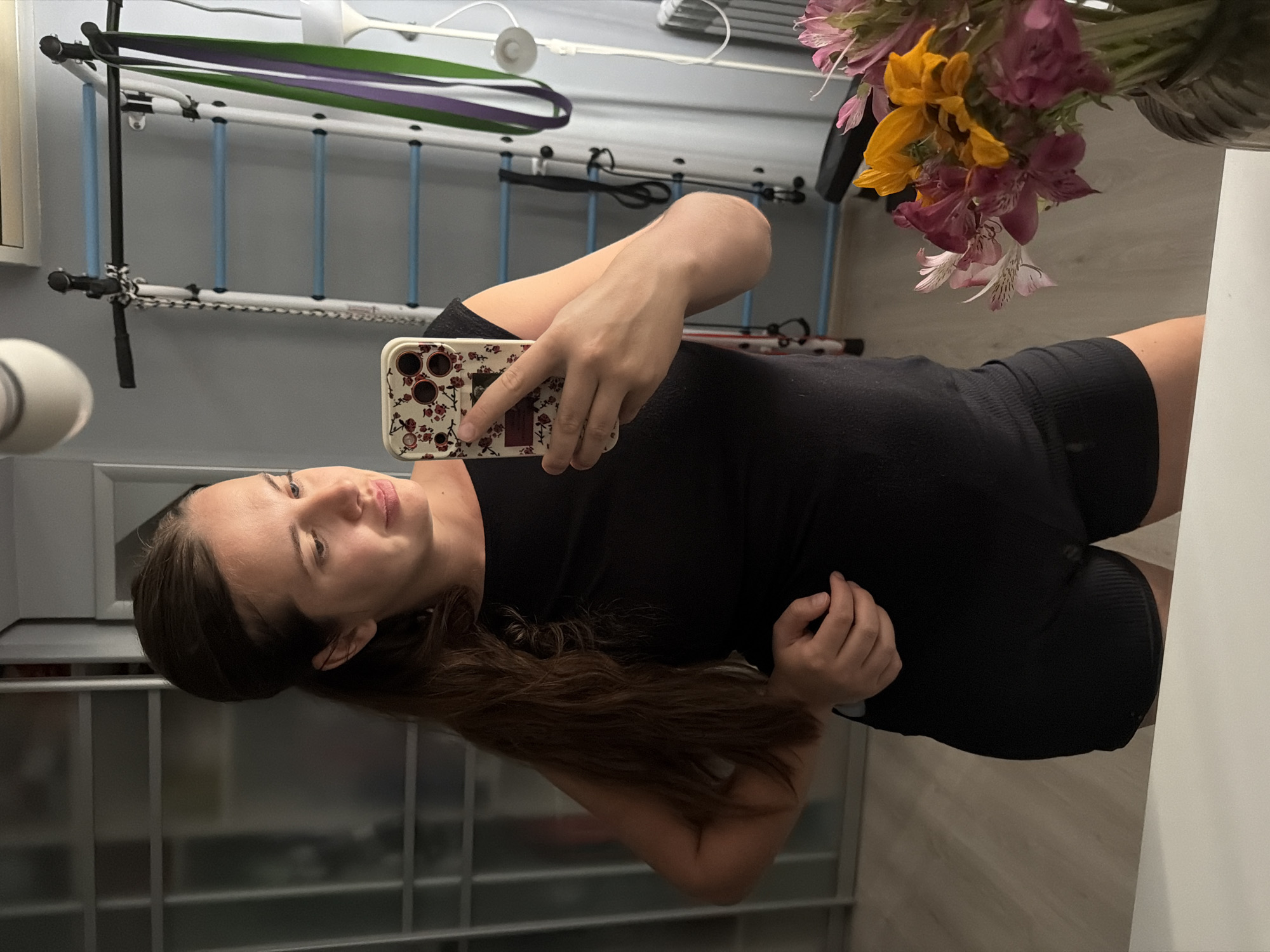 A woman stands in front of a mirror, taking a selfie. She is wearing a black tank top and black shorts. Her hair is brown and pulled back into a ponytail. She is looking at her phone, which has a floral patterned case. In the background, there is a home gym setup with a pull-up bar and resistance bands. There is also a vase of flowers on a table in the foreground. The scene appears to be in a residential setting.