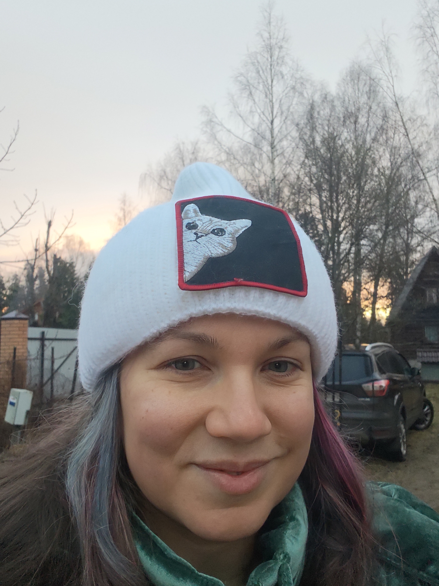 The image shows a woman in a white knitted hat with a patch of a cat on it. She is wearing a green velvet jacket. Her hair has streaks of blue and pink. Behind her, there is a house, trees, and a black SUV parked in a grassy yard. A fence and a brick structure are visible on the left. The sky indicates either sunrise or sunset. The location is Свистуха (кузяевский С/о), Russia.