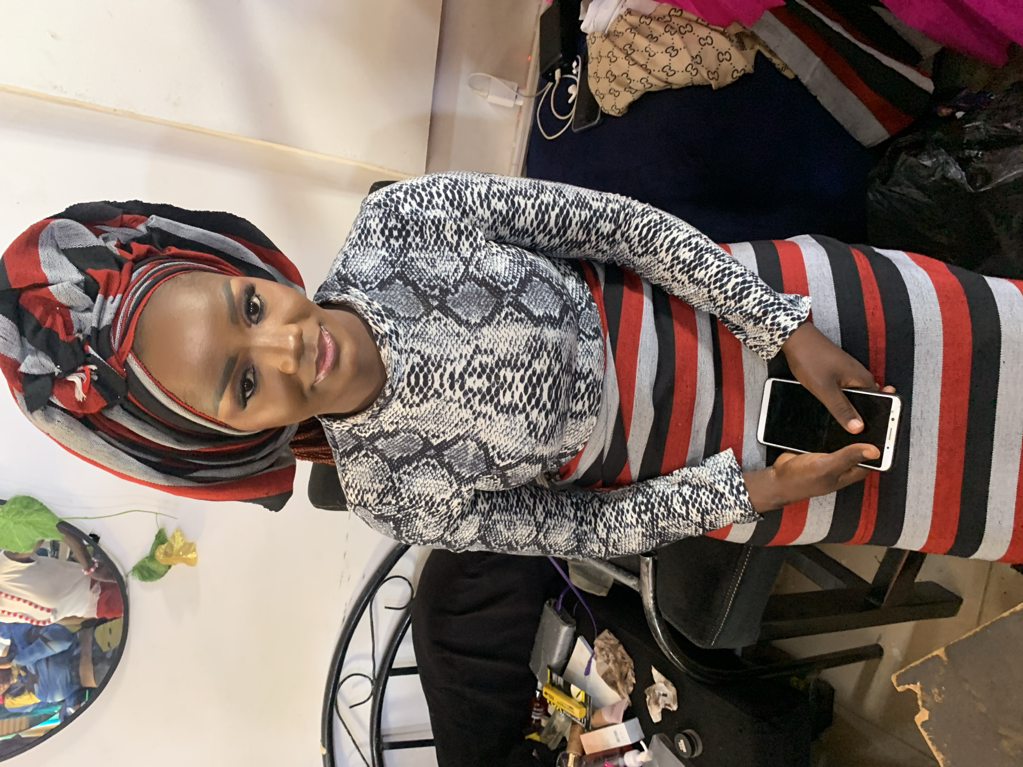 A woman is seated indoors, holding a smartphone. She wears a patterned headwrap and a snakeskin print long-sleeved shirt, and a striped dress. The surrounding environment includes a mirror, chair, and various cosmetics and personal items on a nearby surface. The location is identified as Jos, Nigeria.