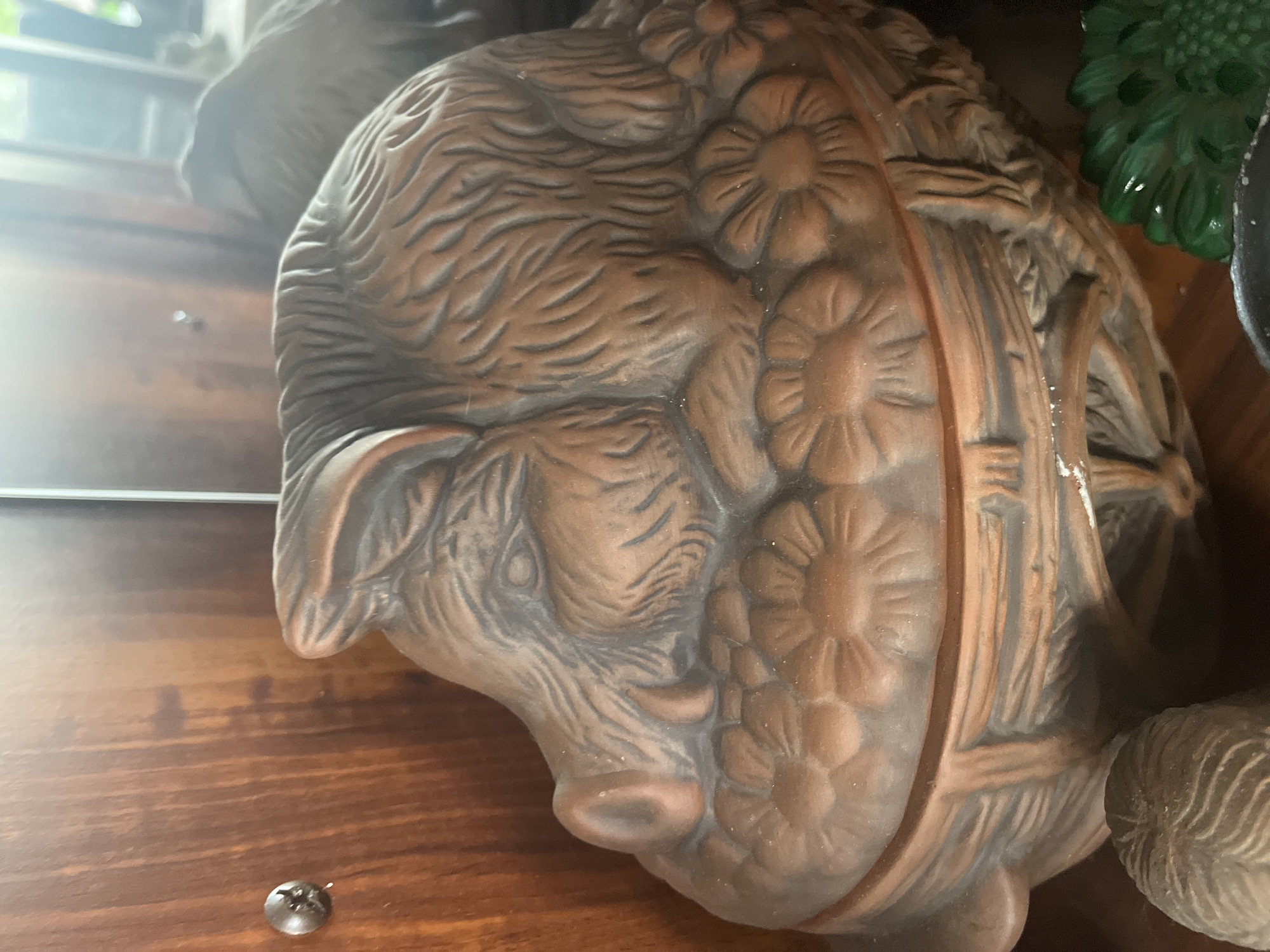 A close-up shot captures a brown ceramic pot shaped like a sleeping boar, its body adorned with textured fur and resting on a bed of carved flowers. The pot itself sits within a wooden cabinet, a metal screw visible on the left side of the frame. The background is blurred, suggesting an indoor environment. The scene is set in the city of Vladimirovka, Russia.
