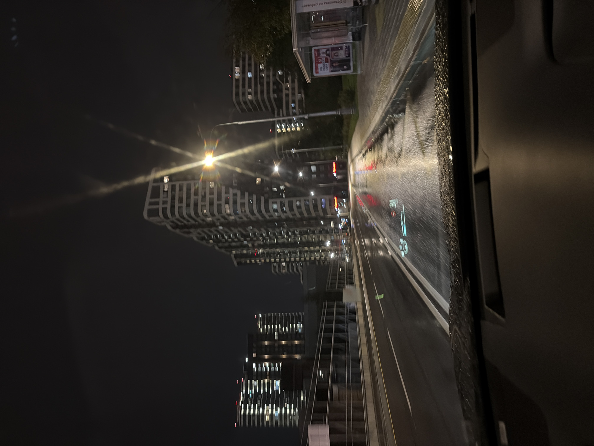 The image shows a view from inside a vehicle at night, looking forward onto a wet city street in Moscow, Russia. The road is slick with rain, reflecting the lights of the surrounding buildings and streetlights. Tall, modern apartment buildings with many illuminated windows line the right side of the street. A bus stop shelter is visible on the sidewalk. The vehicle's dashboard is partially visible in the foreground, and a digital display is projected onto the windshield, showing a speed of 31. The overall atmosphere is dark and rainy, typical of an urban environment at night during inclement weather.