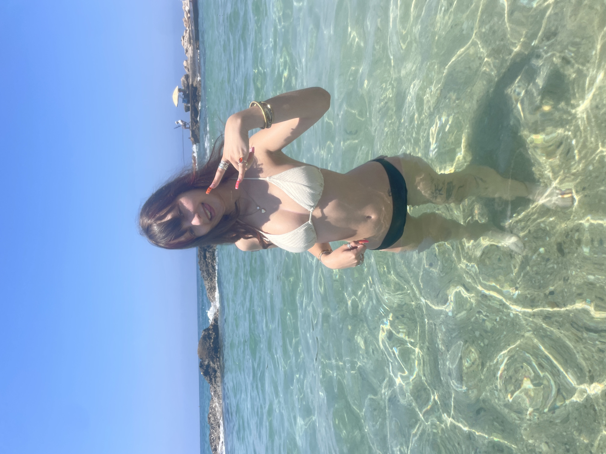 A young woman with dark hair, bangs, and red highlights stands in shallow, clear turquoise water. She is wearing a white crocheted bikini top and black bikini bottoms. Her right arm is raised, and her hand is making a peace sign with her index and middle fingers extended. She is also wearing a ring on her right ring finger and a bracelet on her right wrist. Her left arm is down by her side. She is sticking her tongue out slightly and looking at the camera. The water is clear and shows ripples and the sandy bottom. In the background, there are rocks and a small structure with a yellow umbrella, possibly a lifeguard station, on a rocky outcrop. The sky is a bright, clear blue. The location appears to be a beach in Paphos Municipality, Cyprus, likely along the coastline known for its clear waters and rocky shores.