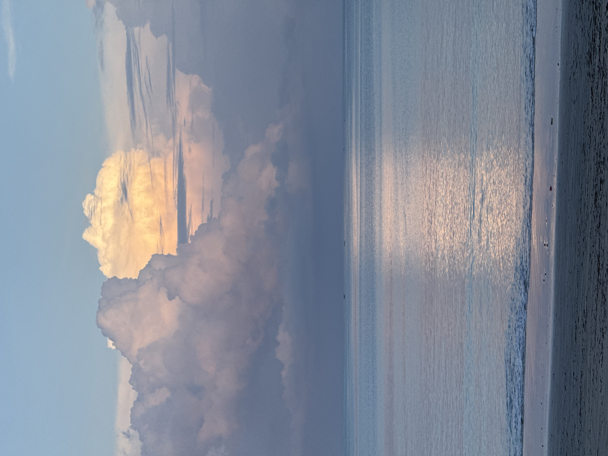The image depicts a serene beach scene, likely taken in Jimbaran, Indonesia, given the location context. Dominating the left side of the frame is a large, cumulonimbus cloud formation with a prominent golden-yellow hue on its upper portion, suggesting sunrise or sunset. The lower part of the cloud is a muted gray-blue, contrasting with the lighter blue sky. The right side of the image features a calm ocean with a glassy surface reflecting the ambient light, creating a shimmering effect. A narrow strip of sandy beach is visible on the far right, marked by gentle waves lapping the shore. A few small, dark figures are standing in the ocean. The overall tone of the image is peaceful and tranquil.