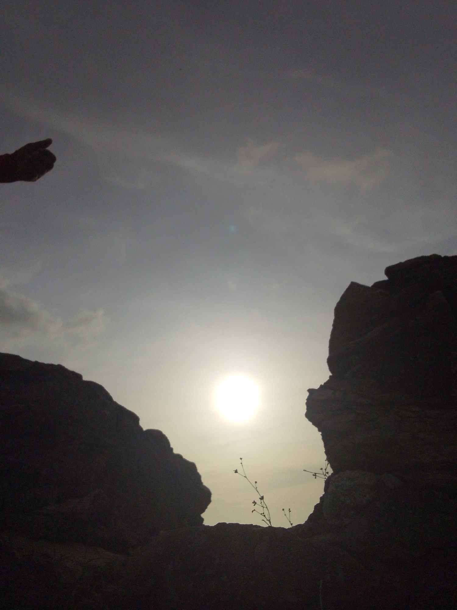 The image depicts a silhouetted view of the landscape near Juan Griego, Venezuela, with a bright sun as the central element. Two large, dark rock formations frame the scene, creating a natural opening. A human hand, also silhouetted, is visible in the upper left corner, with a finger pointing towards the sun. In the foreground, silhouetted against the sky, are thin, dry branches of plants. The sky is a gradient of grey and white, with some wispy clouds. The overall impression is one of looking through a natural archway at the sun.