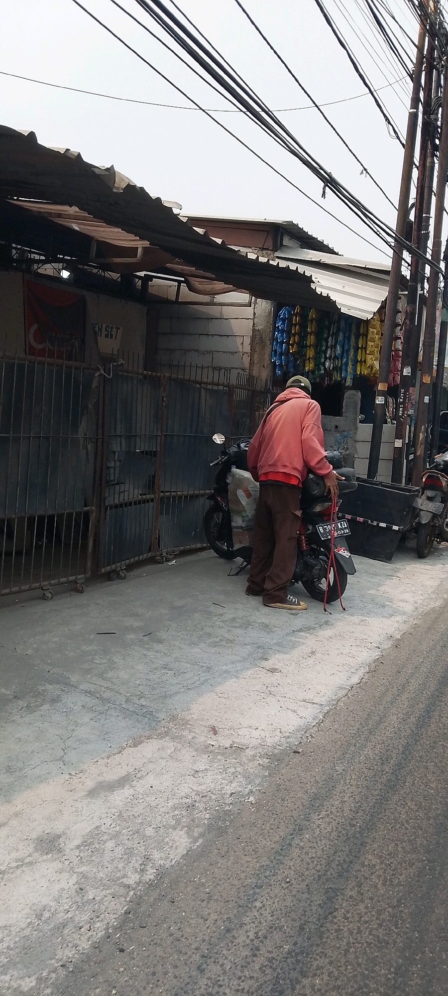In Bekasi, Indonesia, a man wearing a coral-colored hoodie and brown pants stands next to a black scooter. He is leaning over the scooter, with his hands on its handlebars. The scooter has a black box attached to its rear and a red rope is tied to its tail. In the background, a corrugated metal roof extends over a structure with cinder block walls. Several rows of colorful packets, possibly snacks or candies, hang from the side of the structure. A metal gate with vertical bars is situated in front of the building. Overhead, thick electrical wires crisscross the sky. The ground is paved with concrete and asphalt.