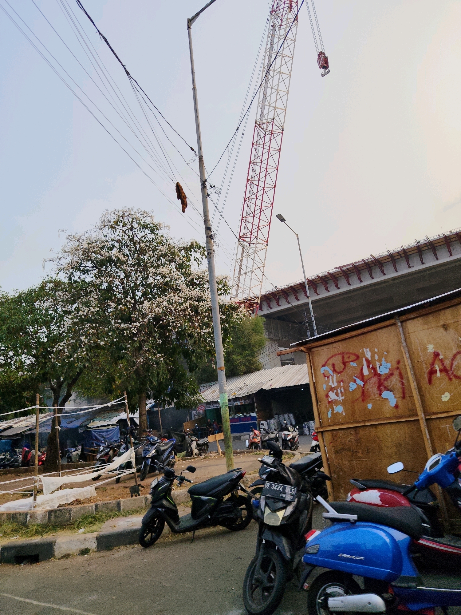 The image depicts a street scene in South Jakarta, Indonesia, with a large construction project underway. A towering red and white crane is visible against the sky, along with a concrete overpass under construction, supported by red metal beams. In the foreground, several parked motorcycles are lined up on the roadside. A light pole with numerous electrical wires stretches across the frame, and a brown, graffitied structure occupies the right side of the image. A blooming tree with white flowers adds a touch of nature to the urban landscape. Tents and market stalls are set up in the background, suggesting a bustling local area. The overall impression is one of ongoing development and daily life in a busy urban environment.