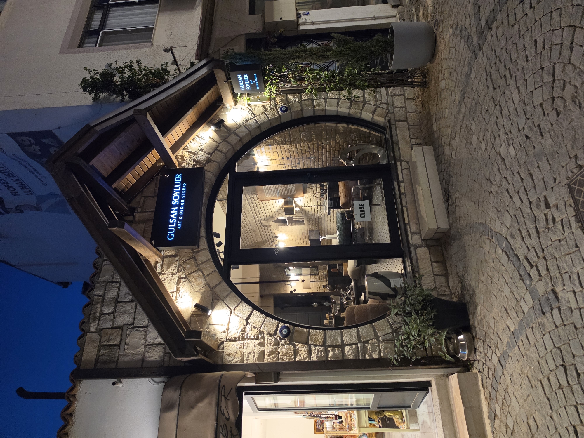 This media file captures the exterior of a shop called "GULSAH SOYLUER ART & DESIGN STUDIO" in Urla, Turkey. The scene is set at night, with artificial lighting illuminating the stone building and cobblestone street. The shop features a distinctive arched entryway with a large glass door and window, offering a glimpse into the interior where furniture and artwork are visible. A neon sign above the entrance displays the shop's name and service. A "CLOSE" sign is visible on the door, indicating the shop is not open. Plants in pots add a touch of greenery to the scene, and a blue "evil eye" charm is mounted on the stone wall. The overall impression is one of a charming, artistic establishment in a European-style setting. There is also visible text on posters on the left side of the image.