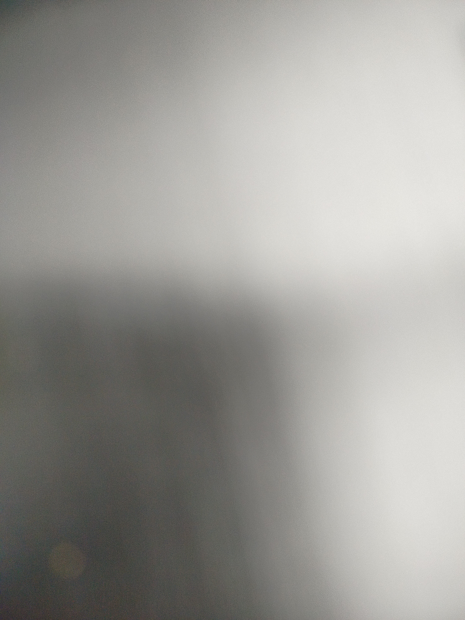 The image is an out-of-focus, grayscale gradient. The upper portion of the image is lighter, gradually transitioning to a darker shade towards the bottom. There is a soft, diffuse circular light anomaly near the lower left corner. There is no discernible information to identify a specific location in Yerevan, Armenia, or any details about people, subjects, objects, actions, interactions, or the environment beyond the gradient and light anomaly.