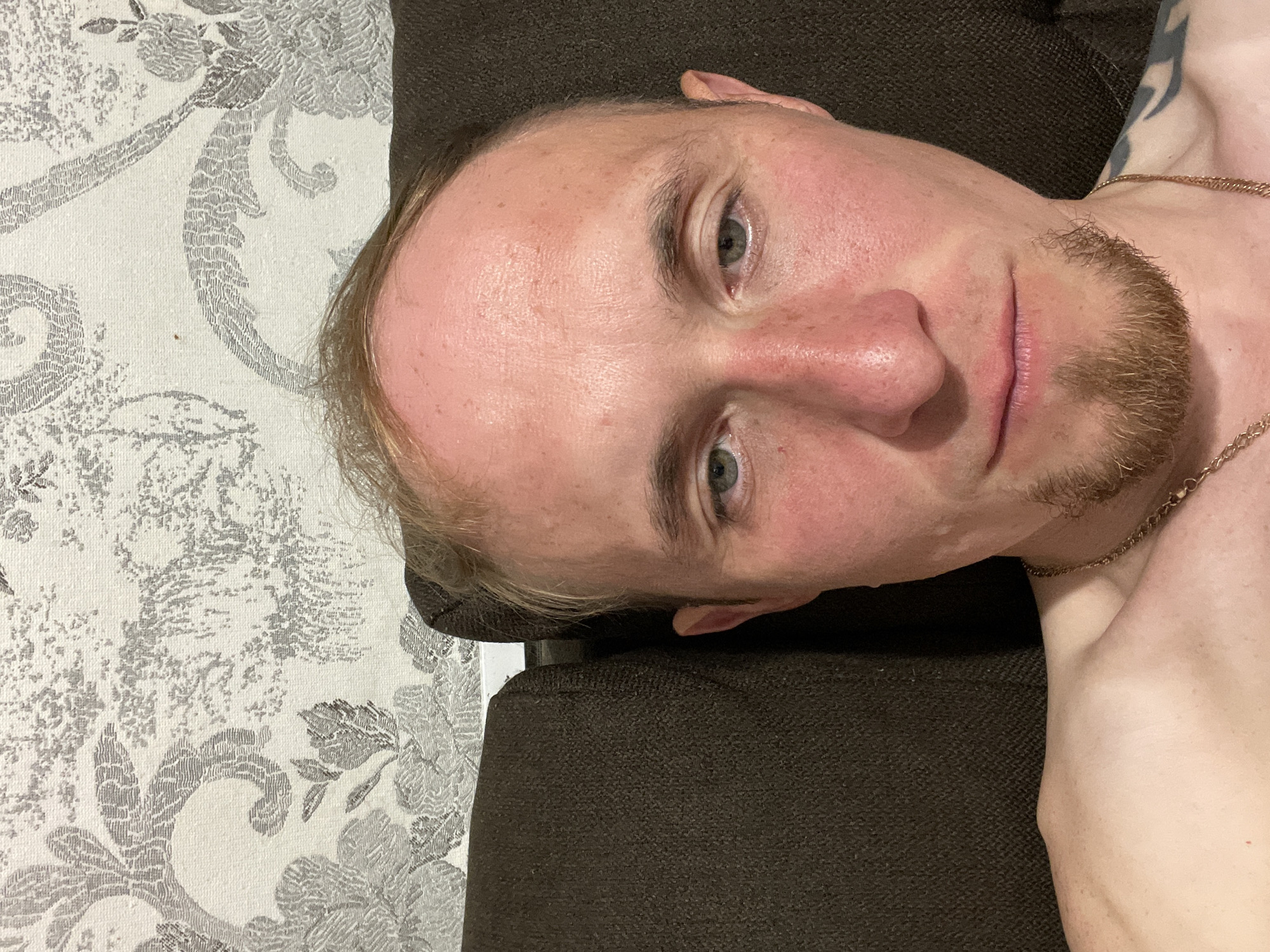 A man is pictured from the chest up, seated against a dark brown couch with a textured, floral-patterned wallpaper in the background. The man has fair skin with visible redness on his cheeks and forehead, a receding hairline with light brown hair, and green eyes. He has a short goatee and is wearing a gold-colored chain necklace. A tattoo is partially visible on his right shoulder. The overall lighting is soft, and the focus is on the man's face. The image is a close-up, likely taken indoors. No specific location in Sarapul, Russia, can be identified from the provided image.