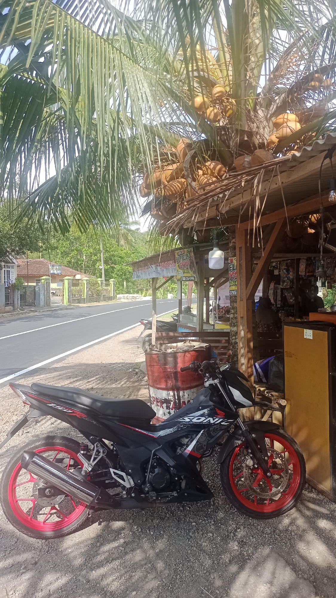 In Cibeber, Indonesia, a black and red motorcycle with red rims is parked on a gravelly shoulder next to a paved road. A makeshift stall with a corrugated metal roof thatched with palm fronds is visible to the right of the motorcycle, displaying various goods and signage in Indonesian. A large, red, oil drum is situated behind the stall. Coconut trees laden with coconuts hang over the stall and partially obscure the sky. The road leads to a residential area with houses visible in the distance, surrounded by lush green vegetation and hills. The scene appears to be a roadside market or convenience store in a rural or semi-rural area of Cibeber.