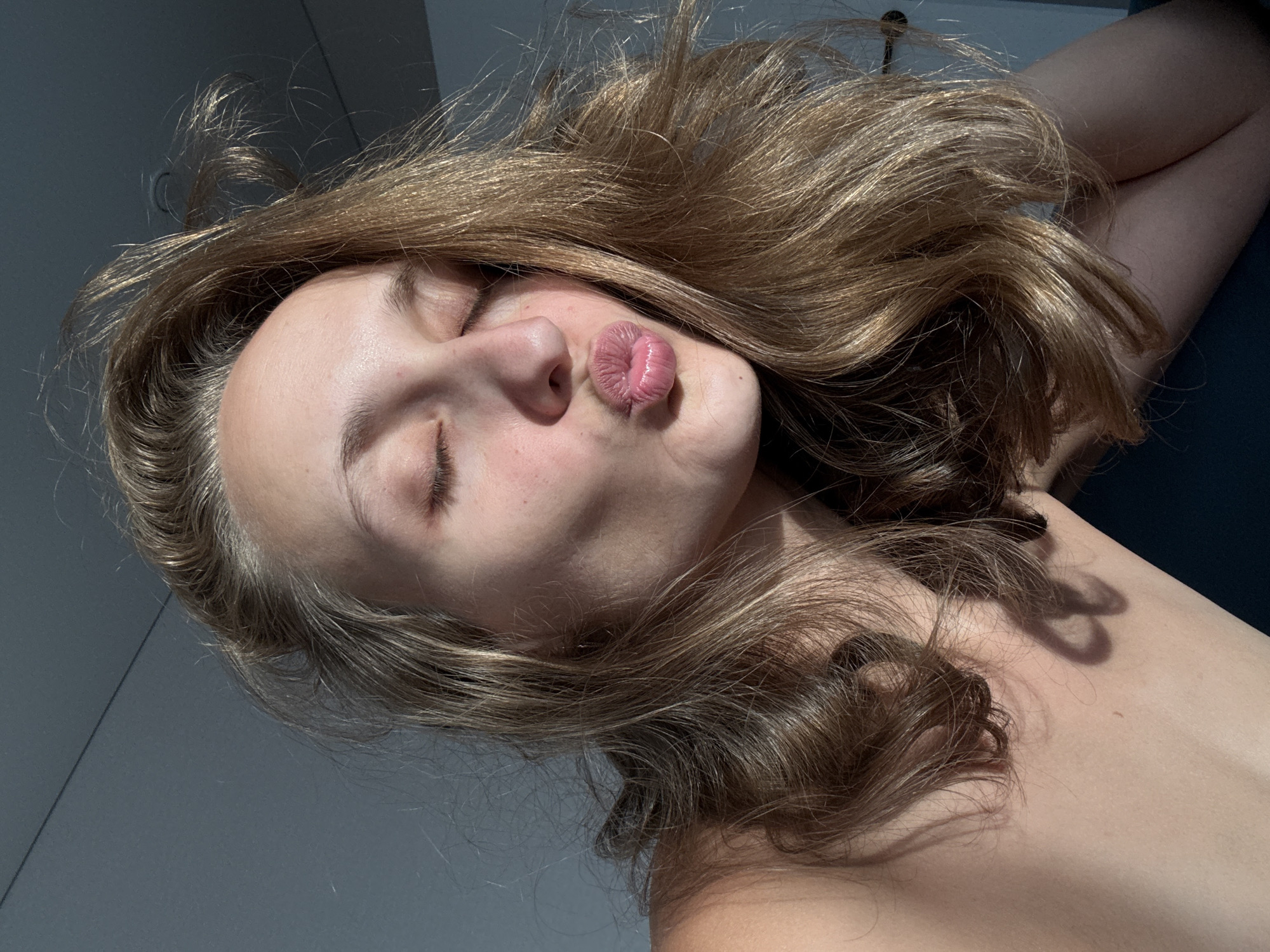 The image is a close-up, low-angle selfie of a young Caucasian woman with her eyes closed and lips puckered in a kiss. She has shoulder-length, wavy blonde hair that is slightly messy and falling around her face. The lighting suggests it's either early morning or late afternoon, with a strong light source coming from the right, casting shadows on her skin. She appears to be indoors, with a plain grey wall in the background. Her bare shoulders are visible. The overall impression is casual and intimate. There is no information available in the image to determine the location in the city of Подшивалово, Russia.