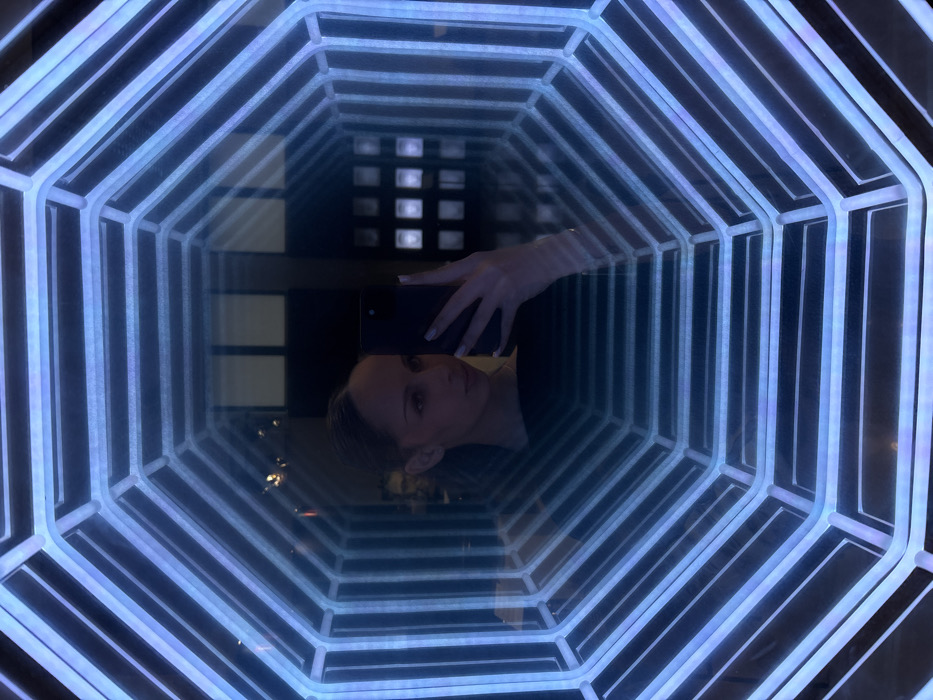 A female is depicted in a mirror selfie taken indoors at The Steps Sofia (Hotel and Co-living space) located at ul. "Lachezar Stanchev" 5, 1756 Sofia, Bulgaria. The subject, visible from the chest up, has light brown hair styled back and holds a black smartphone vertically with her right hand, its back facing the viewer. She is wearing a dark, possibly black, garment. The image is framed by a glowing blue octagonal tunnel, an art installation utilizing mirrors and LED lighting to create an illusion of infinite depth. Faint reflections of an interior background, including rectangular light panels and an indistinct human form, are visible through or within the mirror effect. The environment is artificially lit, dominated by the blue light of the installation.