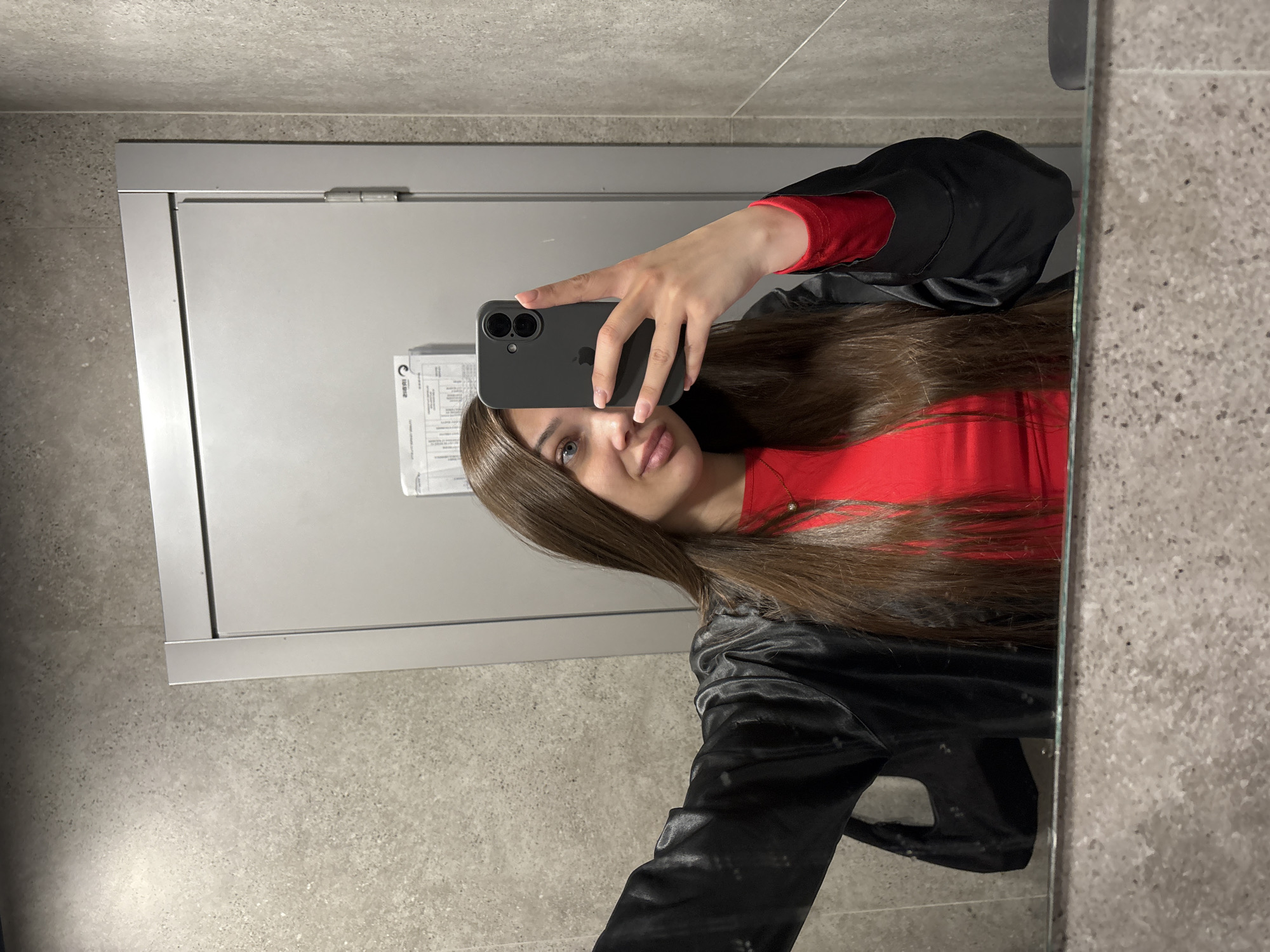 In a restroom with speckled gray walls and a reflective surface, a young woman with long, dark brown hair and light blue eyes takes a selfie in a mirror. She is wearing a red long-sleeved shirt and an open black blazer. Her right hand holds a dark gray smartphone, and her left arm is extended, holding a black handbag. The background features a plain gray door with a silver frame. The scene appears to be located in Kazan, Russia, given the context of the image.