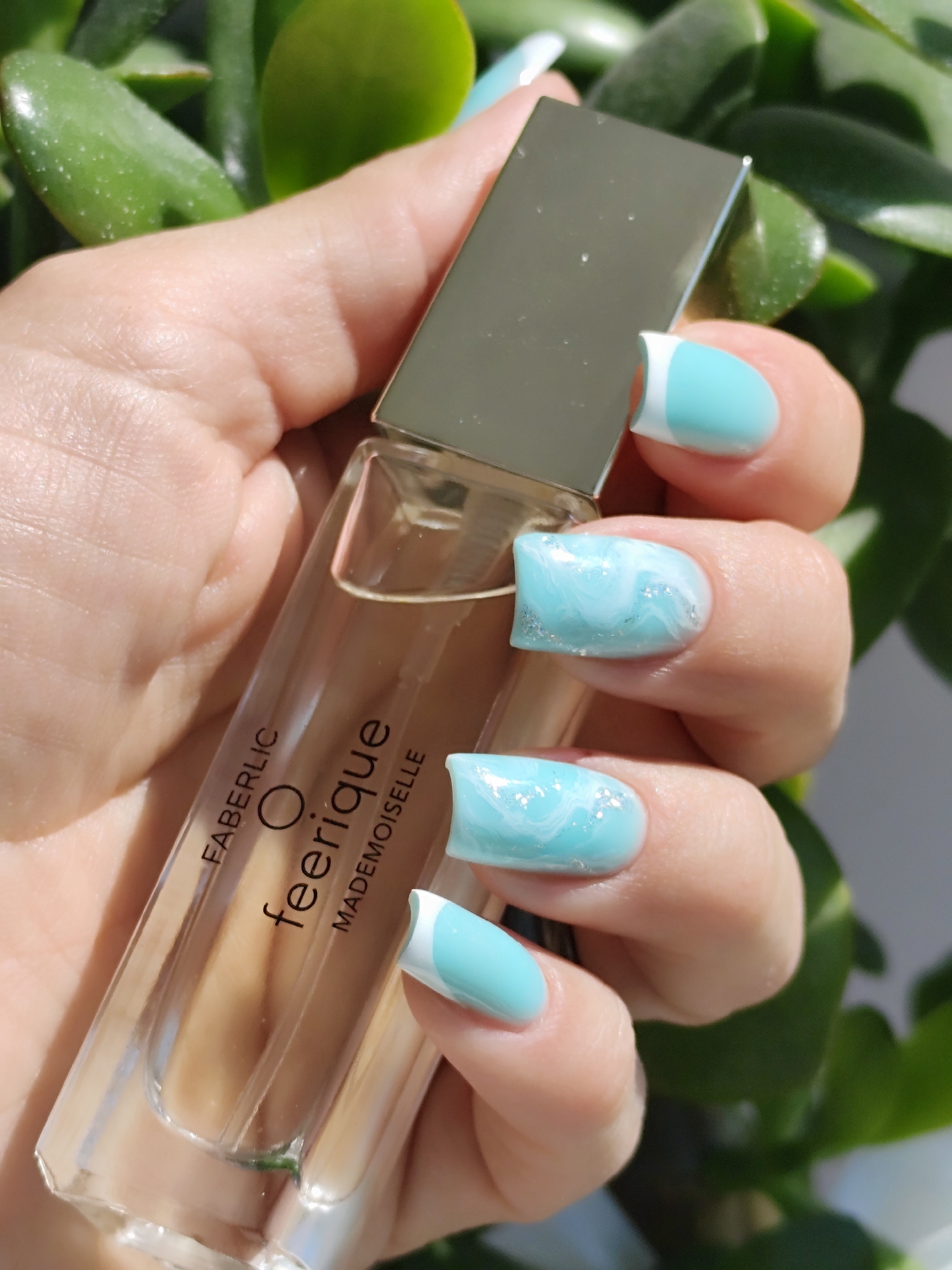 A person holds a perfume bottle in their hand, showcasing a blue-themed nail design with white accents. The perfume bottle is clear with a silver cap, labeled "FABERLIC O feerique MADEMOISELLE". The background includes blurred green foliage. The image was taken in the city of Фоки, Russia.