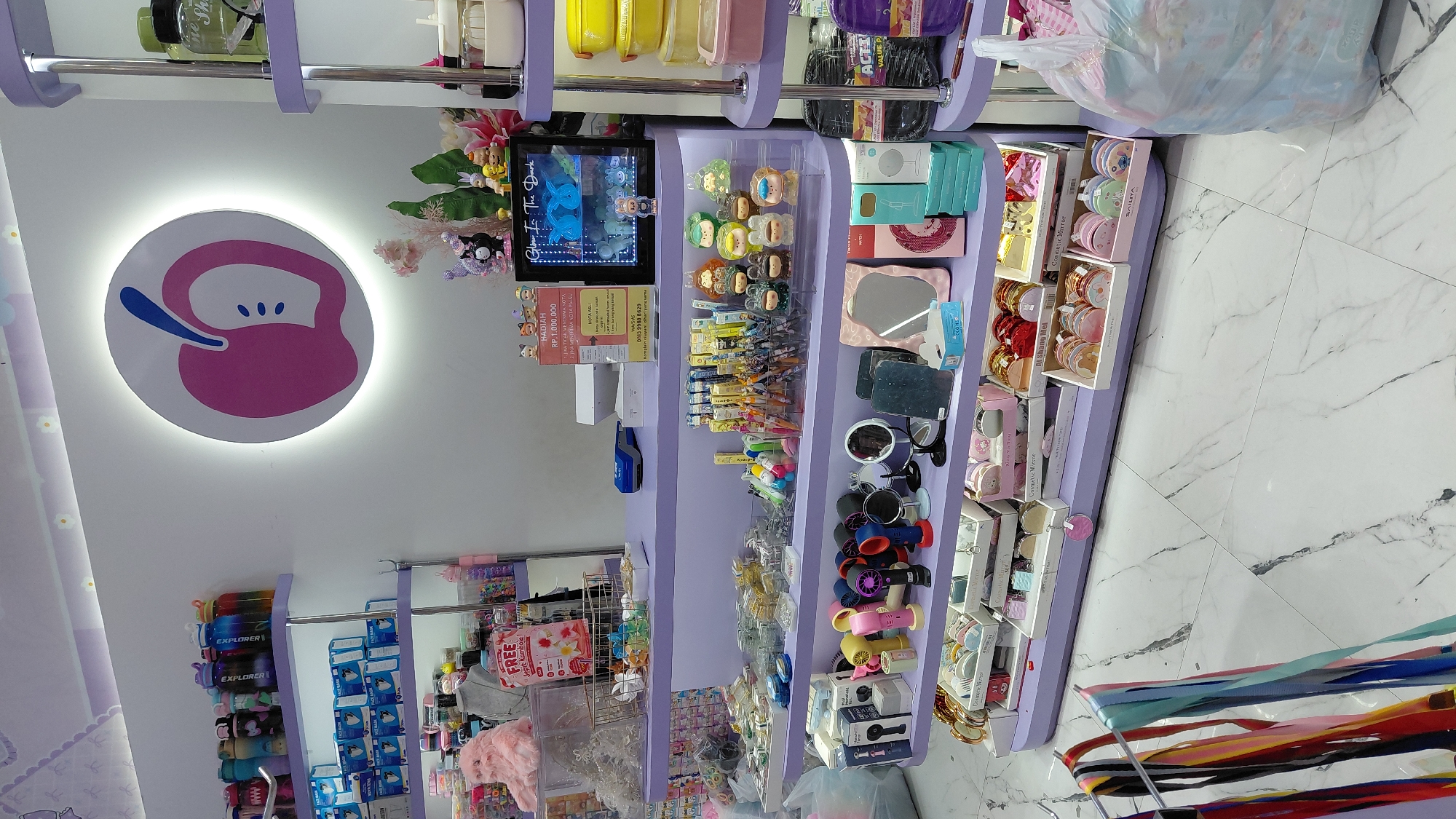 The image shows the interior of a retail store in Purwokerto, Indonesia, specializing in cute and whimsical items. The store features light purple shelving units filled with various products. Prominently displayed is a large, illuminated circular sign with a pink apple motif on the white wall.
On the top shelf, an array of colorful water bottles with blue and white labels are arranged. Below, a pink plush cat sits atop a clear display case. A red and white promotional sign, likely advertising a "FREE JELE KEMUBAU" offer, is also visible on this shelf.
Further down, shelves are packed with small merchandise. One shelf holds an assortment of hair accessories and novelty items, including small figurines of characters and animals. Another shelf displays compact mirrors, small handheld fans in various colors, and other cosmetic accessories. The lowest visible shelf contains more decorative items and what appear to be sets of small boxes.
In the background, a counter area is set up with a blue card reader and a stack of white paper. To the right of the counter, a lit display case contains blue, glowing figurines. A sign above this case reads "Glow In The Dark".
The floor is tiled with a white marble pattern. To the left of the shelves, colorful ribbons hang from a display rack. A large, translucent plastic bag filled with what appears to be textiles or plush items is on the floor to the right. The overall aesthetic of the store is bright, feminine, and playful, suggesting it caters to a younger demographic or those interested in kawaii culture.