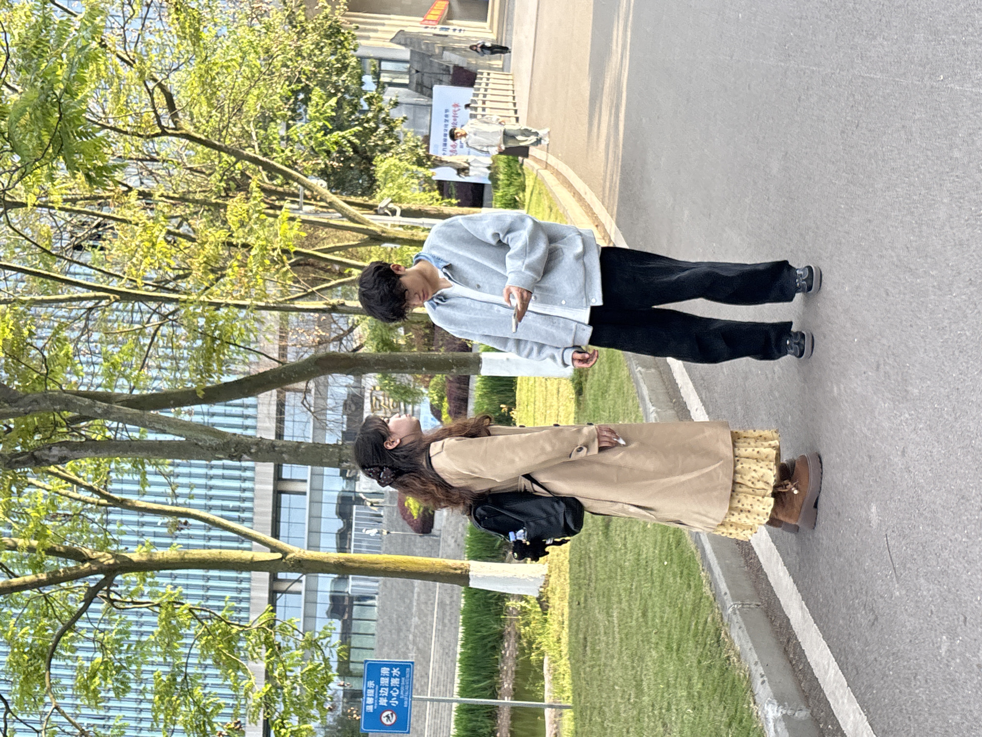 A young man and woman stand on a paved walkway, likely within a park or campus in Jiangjin, China. The man, facing to the right, is dressed in a gray jacket and black pants, with his attention focused on a handheld device, possibly a mobile phone. The woman, standing to his left and facing toward the sky, wears a tan trench coat over a yellow patterned dress, paired with brown boots and a black backpack. Trees with budding green leaves flank the walkway. A modern building with glass and steel construction is visible in the background, behind the woman. A blue sign with Chinese characters stands near the walkway's edge.