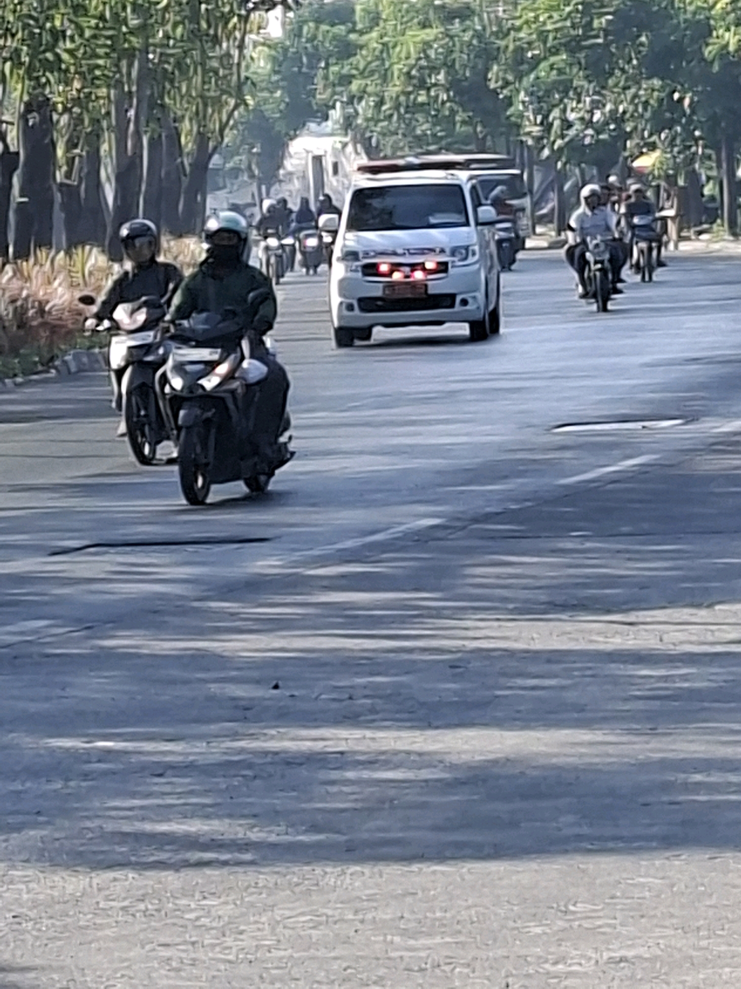 In an outdoor daytime scene on a paved road in Surabaya, Indonesia, two individuals ride on a black motorcycle in the foreground, with the rider in front wearing a blue helmet and the rider behind wearing a black face mask and a dark green jacket. Behind them, another black motorcycle carries a person with a white helmet and light-colored shirt. Further down the road, a white vehicle with flashing red and blue lights, possibly an emergency vehicle, is visible. The road is lined with trees and some buildings are present in the background. The lighting suggests it is either early morning or late afternoon.