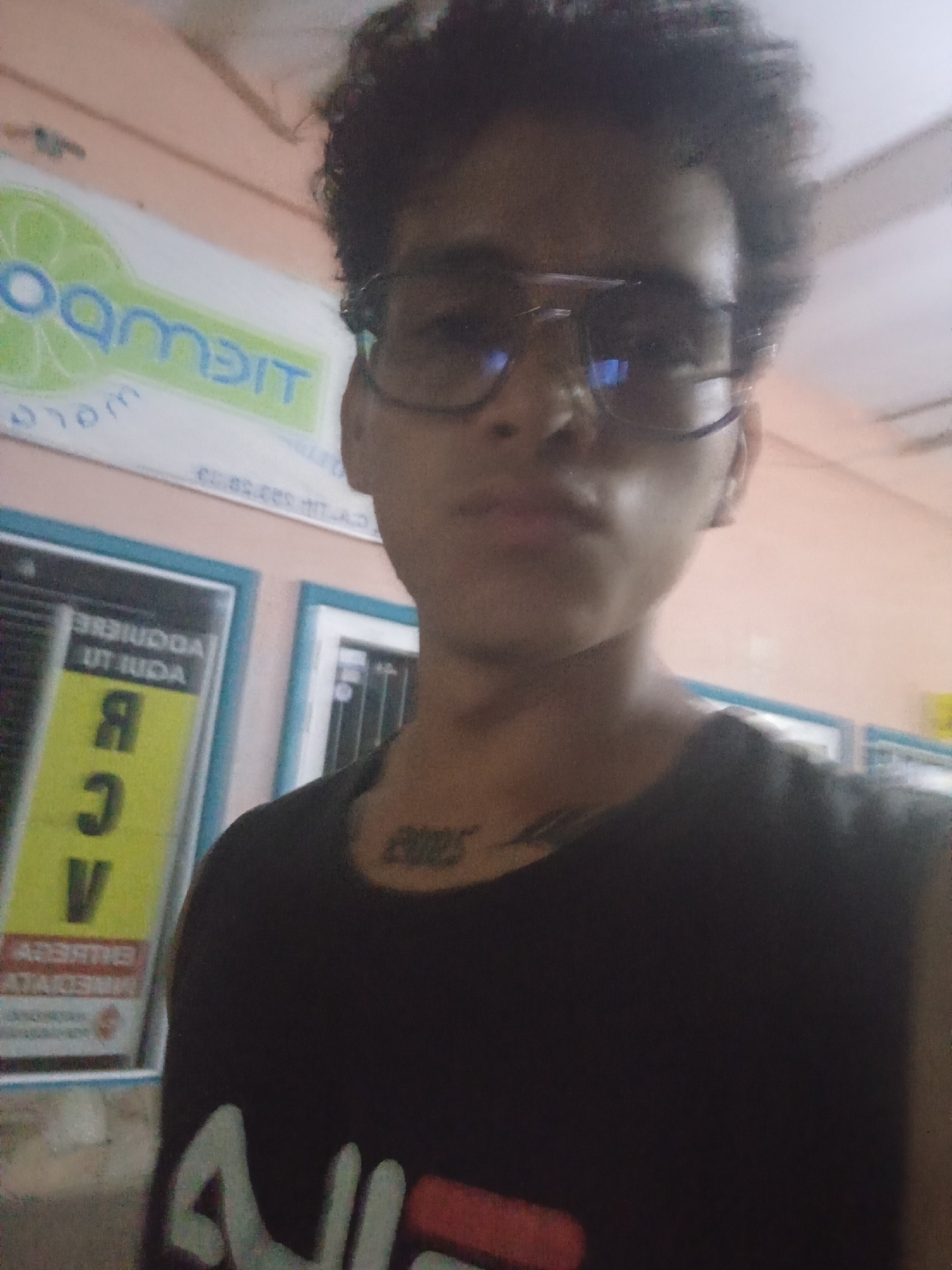A young person with dark, curly hair and glasses is taking a selfie in what appears to be an indoor setting. The person is wearing a black t-shirt with a logo on the chest and a larger logo on the left side of the shirt. The background shows a pink wall with a sign that has blue lettering. To the left of the person, there is a framed poster with yellow and black text. The overall lighting is somewhat dim. The environment suggests a commercial or public space, possibly a shop or an office, in Juan Griego, Venezuela.