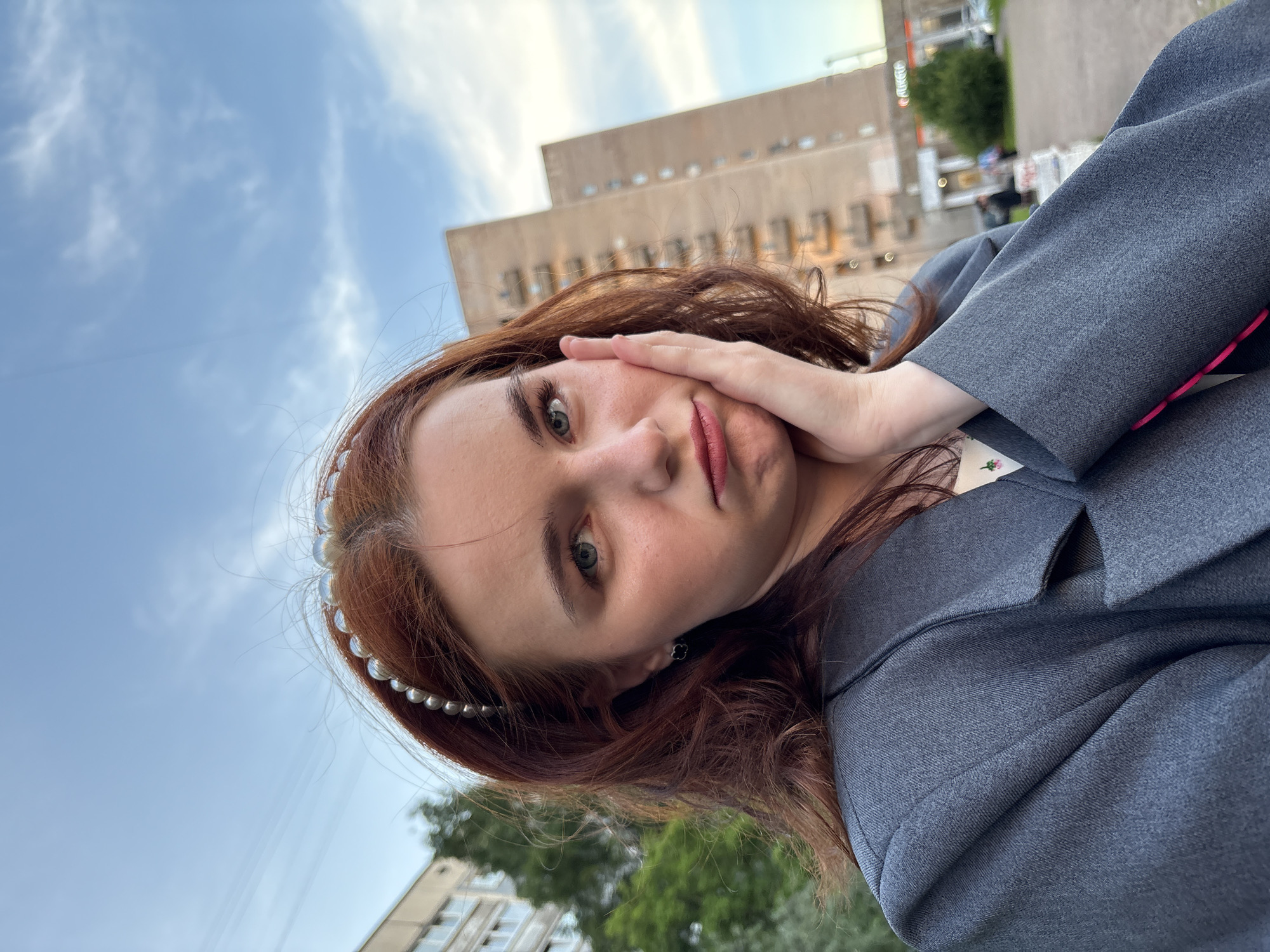 The image is a portrait of a young woman, taken from a low angle and slightly tilted. The primary subject has reddish-brown, wavy hair adorned with a pearl-studded headband. She has light-colored eyes (appearing green or blue) and is wearing red lipstick. Her left hand is positioned against her cheek, gently pressing her face. She wears a dark grey blazer or jacket over a light-colored top with a subtle floral print, visible near her collar. Her expression is direct, looking towards the camera, suggesting a posed selfie or photograph.
The scene is an urban outdoor setting. In the background, a clear blue sky with scattered white clouds dominates the upper half of the image, indicating daytime. Below the sky, several tall, rectangular buildings with numerous windows are visible, characteristic of post-Soviet architecture found in cities like Saint Petersburg, Russia. Some green foliage, likely from trees or bushes, is present in the lower corners of the background. Overhead lines or cables can also be seen against the sky. The lighting suggests a pleasant day, possibly late afternoon. There is no visible text.