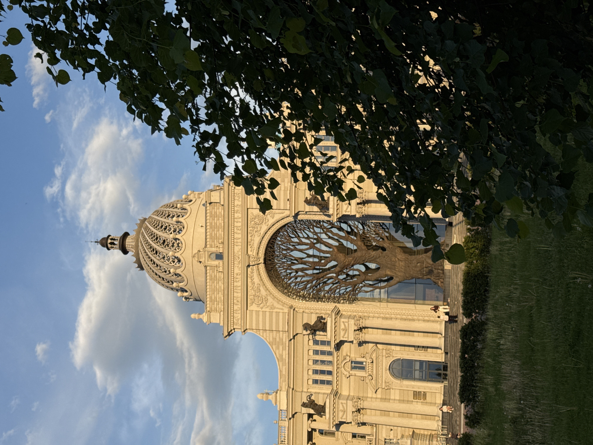 The image features the Palace of Farmers in Kazan, Russia. The building's facade is ornate and constructed in a light-colored stone. A metal tree sculpture is prominently displayed within a large archway. A dome with latticework details sits atop the building. The sky is visible in the background, with some clouds present. Green foliage from a tree frames the top of the image. In the lower right of the image there are steps descending into a grassy area and several people are seen standing on the steps.