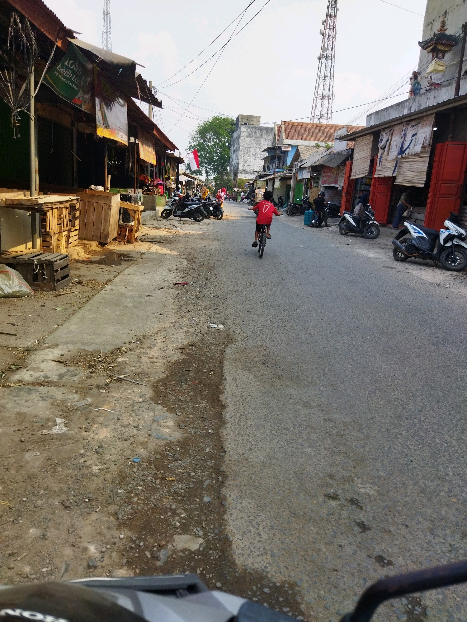 In Banjar Agung, Indonesia, a boy rides a bicycle down a paved road lined with market stalls. The boy, wearing a red shirt with the number 17 on the back, is the central subject. He is positioned on the right side of the road, moving away from the camera. On the left side of the road, wooden market stalls are visible, some with corrugated metal roofs. A white tarp is draped over one stall. Several motorcycles are parked along the right side of the road near the stalls. The environment appears to be a bustling street market, with a clear sky overhead and a telecommunications tower visible in the distance. The ground is a mix of asphalt and concrete, with some debris and vegetation present. The scene captures a moment of everyday life in a local Indonesian community.