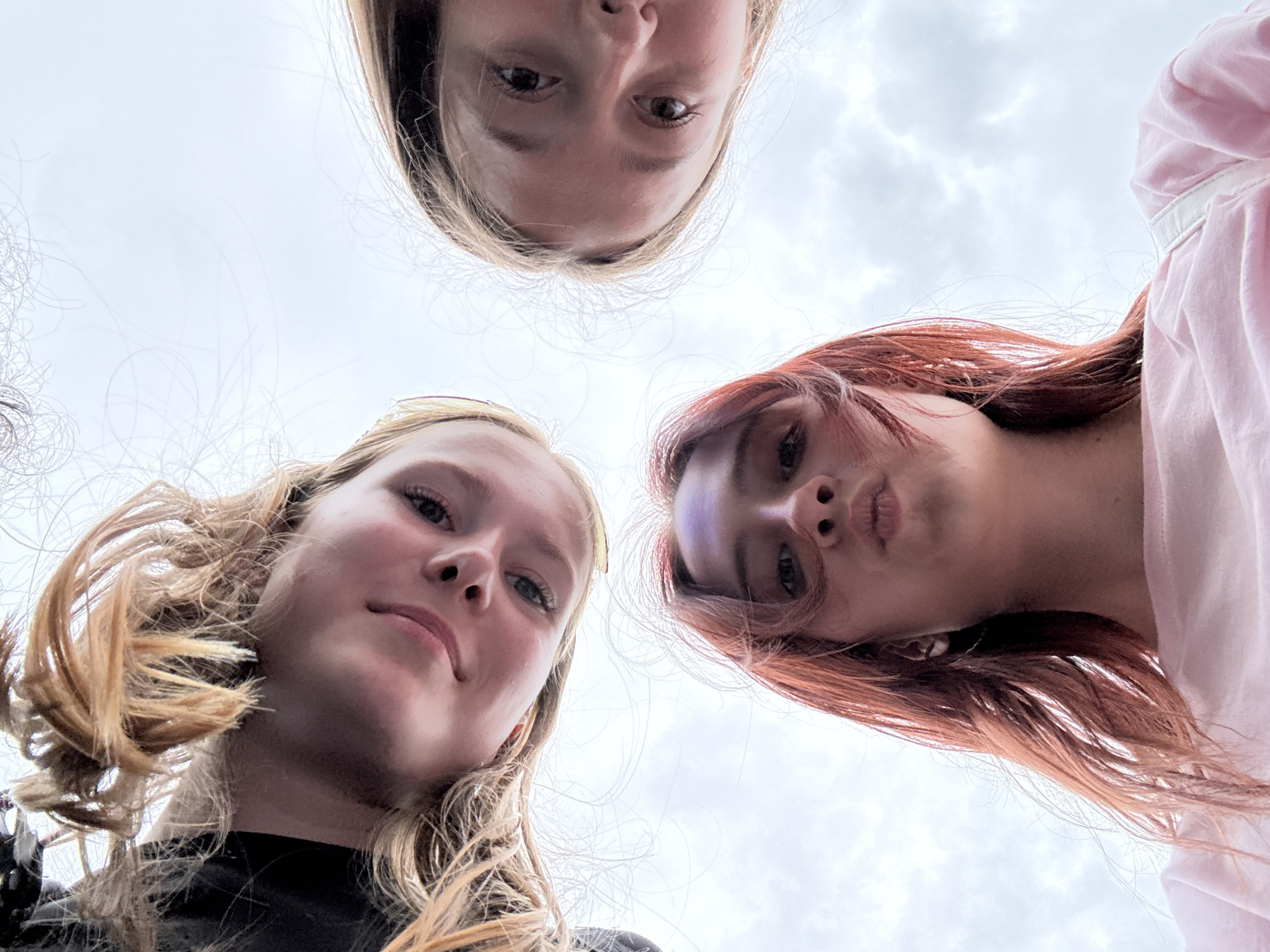 A low-angle, wide-shot photograph captures three young women, appearing to be in their late teens or early twenties, as they look down towards the camera. The sky, with scattered clouds, serves as the backdrop. The women are positioned in a triangular formation, with one in the foreground and two further back and to the sides.
The woman in the foreground, centered in the frame, has reddish-brown hair and is making a "pouty" face, with her lips pursed. She is wearing a light pink t-shirt. A silver earring is visible in her left ear.
To the upper left, another woman with blonde hair is visible, her head tilted and hair falling around her face. She appears to be smiling subtly.
To the upper right, a third woman, also with blonde hair, looks towards the camera with a neutral expression. Her hair is also falling around her face.
The perspective suggests the photo was taken from a low vantage point, possibly from someone lying on the ground or looking up at the women. The overall atmosphere is casual and informal. There is no discernible information in the image to indicate the specific location within the city of Izhevsk, Russia.