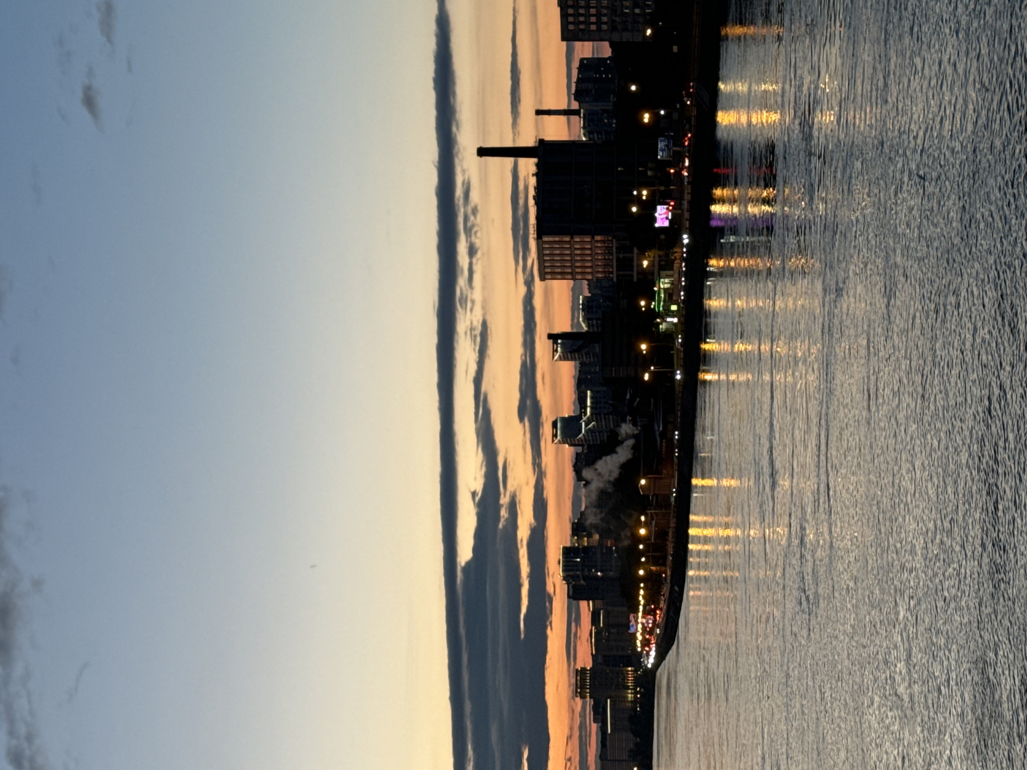 The image captures a panoramic view of the city of Kudrovo, Russia, at dusk. The sky is a gradient of colors, transitioning from a pale yellow at the horizon to a soft blue towards the top, with scattered dark clouds creating a dramatic effect. A prominent cityscape lines the waterfront, featuring a mix of modern buildings and industrial structures, including a distinctive smokestack emitting white vapor, possibly from a power plant or factory. Lights from buildings and streetlamps illuminate the scene, casting reflections on the rippling surface of the wide body of water in the foreground. The water appears calm, with the reflections of the city lights creating streaks of yellow and orange. A road with traffic is visible along the waterfront, indicated by the headlights and taillights of vehicles. The overall atmosphere is serene yet dynamic, showcasing the intersection of urban development and natural elements during the twilight hours.