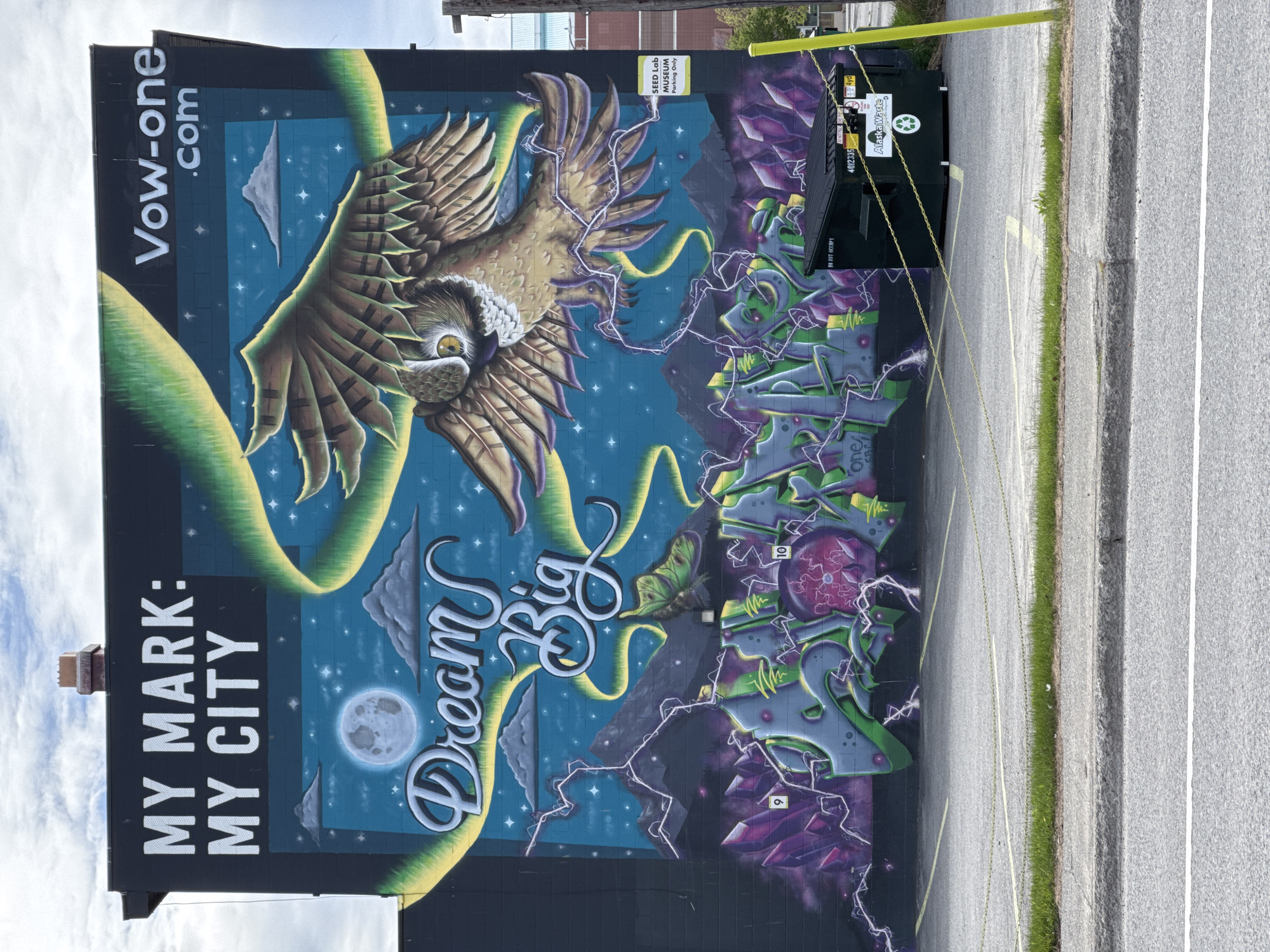 The image displays a vibrant, large-scale mural painted on the side of a dark building, set against a backdrop of a clear, cloudy sky overhead. The mural's central feature is a majestic owl with striking yellow eyes and brown and gold plumage, soaring with outstretched wings against a deep blue night sky filled with white stars and scattered clouds. A prominent full moon is depicted towards the lower left of the mural. Yellow-green ribbons gracefully twine around the owl and across the starry expanse.
Text incorporated into the mural includes "MY MARK: MY CITY" in bold white letters on the left side, and "Dream Big" in elegant script-like font in the center. Vertically oriented text "Vow-one.com" is visible in the upper left corner. The right side of the mural features abstract, geometric designs resembling jagged, purple crystals and green, blocky structures, interconnected by white lightning bolts. Small numerical details, such as "9" and "10," appear on some of these shapes. Another small text "one/CBC" is partially visible at the bottom right of the mural.
In the foreground, a dark green dumpster is placed directly in front of the mural, secured by bright yellow chains. Text on the dumpster reads "Alaska Waste," "4yd," "4012335," and "DO NOT OCCUPY." A sign above the dumpster states "SEED Lab MUSEUM Parking Only." The ground consists of an asphalt parking lot or road, marked with white and yellow lines, and bordered by a strip of green grass and a concrete curb. The scene is illuminated by daylight, providing a clear view of the detailed artwork in Anchorage, United States of America.