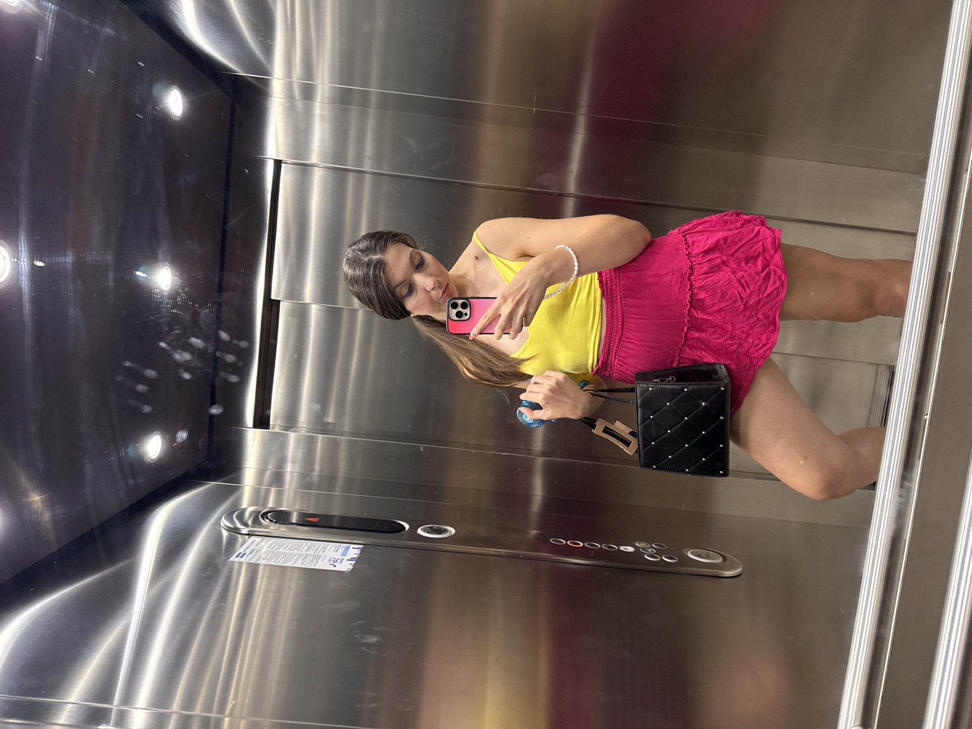 A woman stands in an elevator, taking a selfie. She is wearing a yellow tank top and a bright pink skirt, and is holding a black purse and a blue object. The elevator walls are made of polished metal.