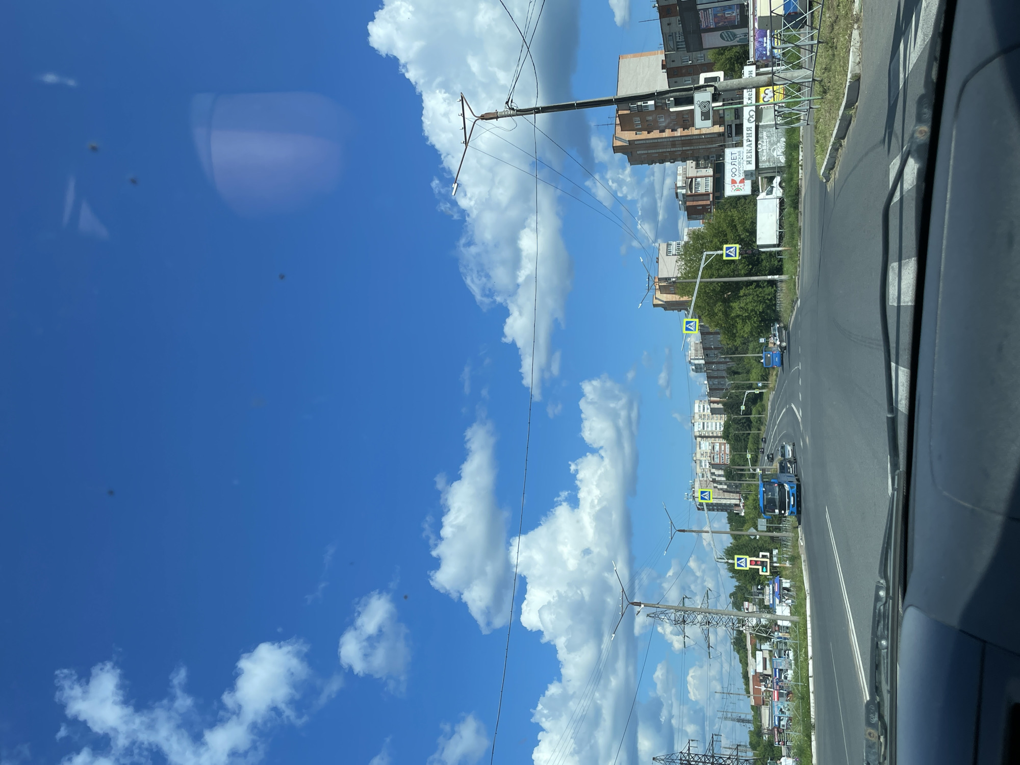 This image captures a view from the driver's perspective through a car's windshield on a clear, sunny day in Kirovo-Chepetsk, Russia. The sky is a vibrant blue with scattered white cumulus clouds. The foreground shows the dashboard and windshield wipers of the vehicle.
The scene depicts a multi-lane road with white lane markings. On the left side of the road, there are power lines supported by tall metal pylons and some low-rise buildings. Further down the road, on the right side, are several multi-story apartment buildings constructed with brick and concrete, characteristic of Soviet-era architecture, interspersed with trees and greenery.
Traffic signs, including pedestrian crossing signs with yellow triangular markers and a traffic light showing red, are visible. A blue bus is in motion on the road, along with other vehicles. Billboards with Russian text are present along the roadside, some advertising a bakery. The overall environment appears to be a suburban or residential area within the city.