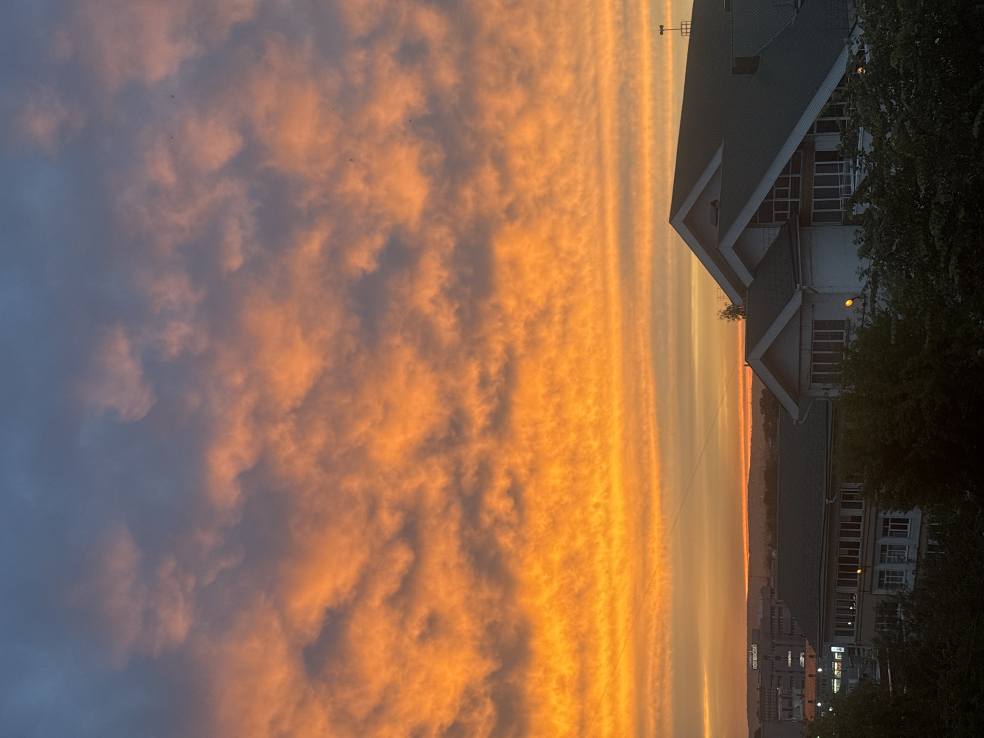 The image captures a vibrant sunset over Irkutsk, Russia. The sky is dominated by dramatic orange and yellow clouds, tinged with grey and blue at the top, creating a striking contrast. Below the fiery sky, the silhouette of a city skyline is visible, with buildings and trees partially obscuring the view. In the foreground, the roofs of buildings are prominent, with one structure featuring a green, sloped roof and a white facade. The overall scene is a picturesque representation of an evening sky in a cityscape.