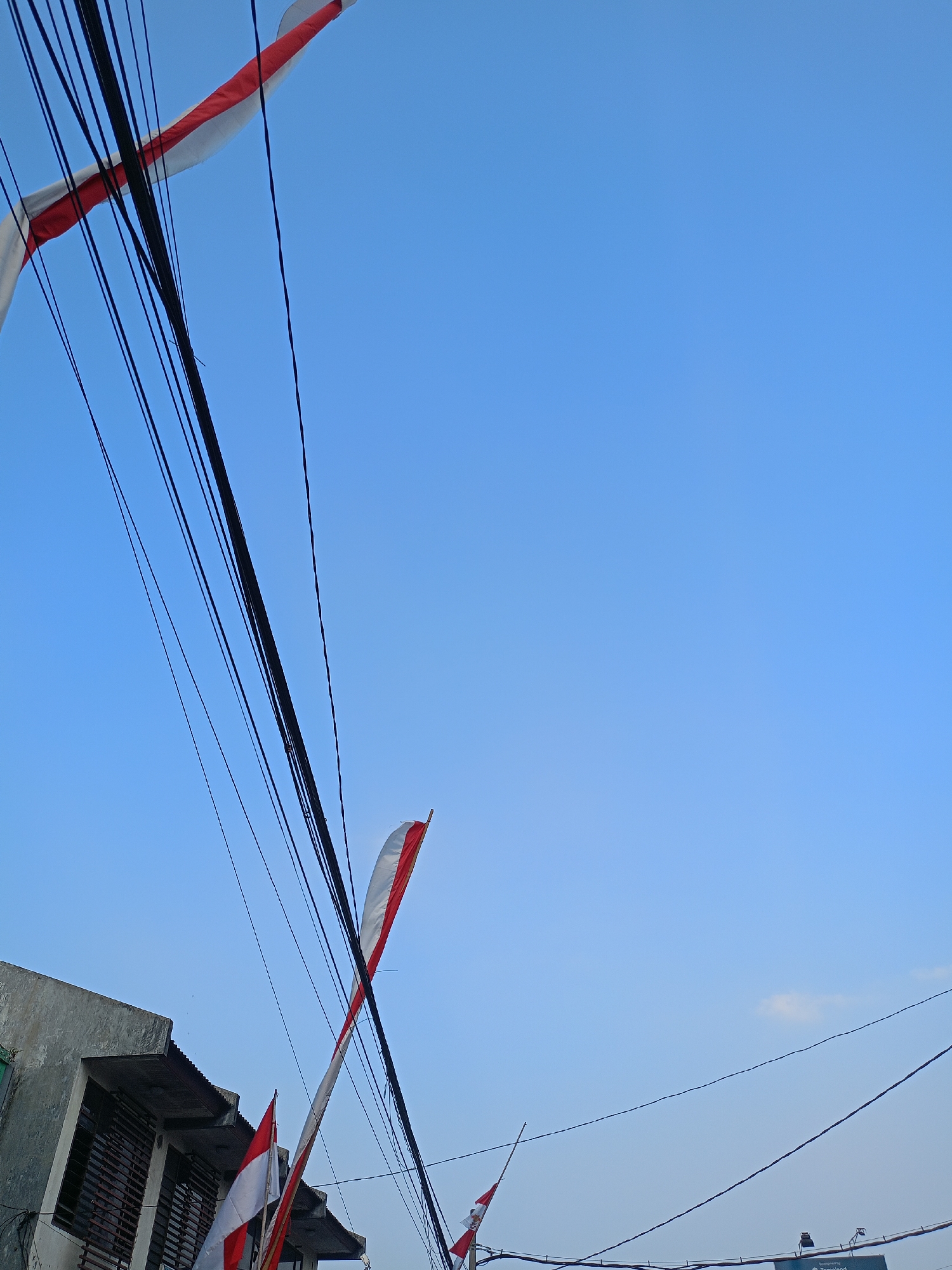 The image depicts a view from below, looking up at a clear blue sky with several Indonesian flags displayed. The flags, with their characteristic red and white horizontal stripes, are mounted on poles and appear to be part of a celebration or commemoration. They are positioned against a backdrop of tangled electrical wires, which dominate the left and upper portions of the frame.
In the lower left corner, a portion of a building is visible. It's a simple structure with a gray, somewhat weathered facade and a corrugated metal roof. The windows have horizontal slats, suggesting they may be for ventilation or security. The building appears to be an ordinary urban or suburban dwelling.
The flags are the most prominent subjects. One large flag is diagonally positioned across the upper left of the image, its red and white colors clearly visible. Another, smaller flag is seen further down and to the left, near the building. A third flag, partially visible, is in the lower right quadrant of the image. These flags are likely displayed in anticipation of or during Independence Day celebrations in Indonesia.
The scene suggests a residential or commercial area within a city. Given the presence of Indonesian flags and the general atmosphere, it's plausible this photograph was taken in Batu, Indonesia, a city known for its natural beauty and cultural events, especially around national holidays. The image captures a moment of patriotic display amidst the everyday infrastructure of electrical wires and local architecture. There are no people clearly visible in the image, the focus being on the flags, wires, and the building.