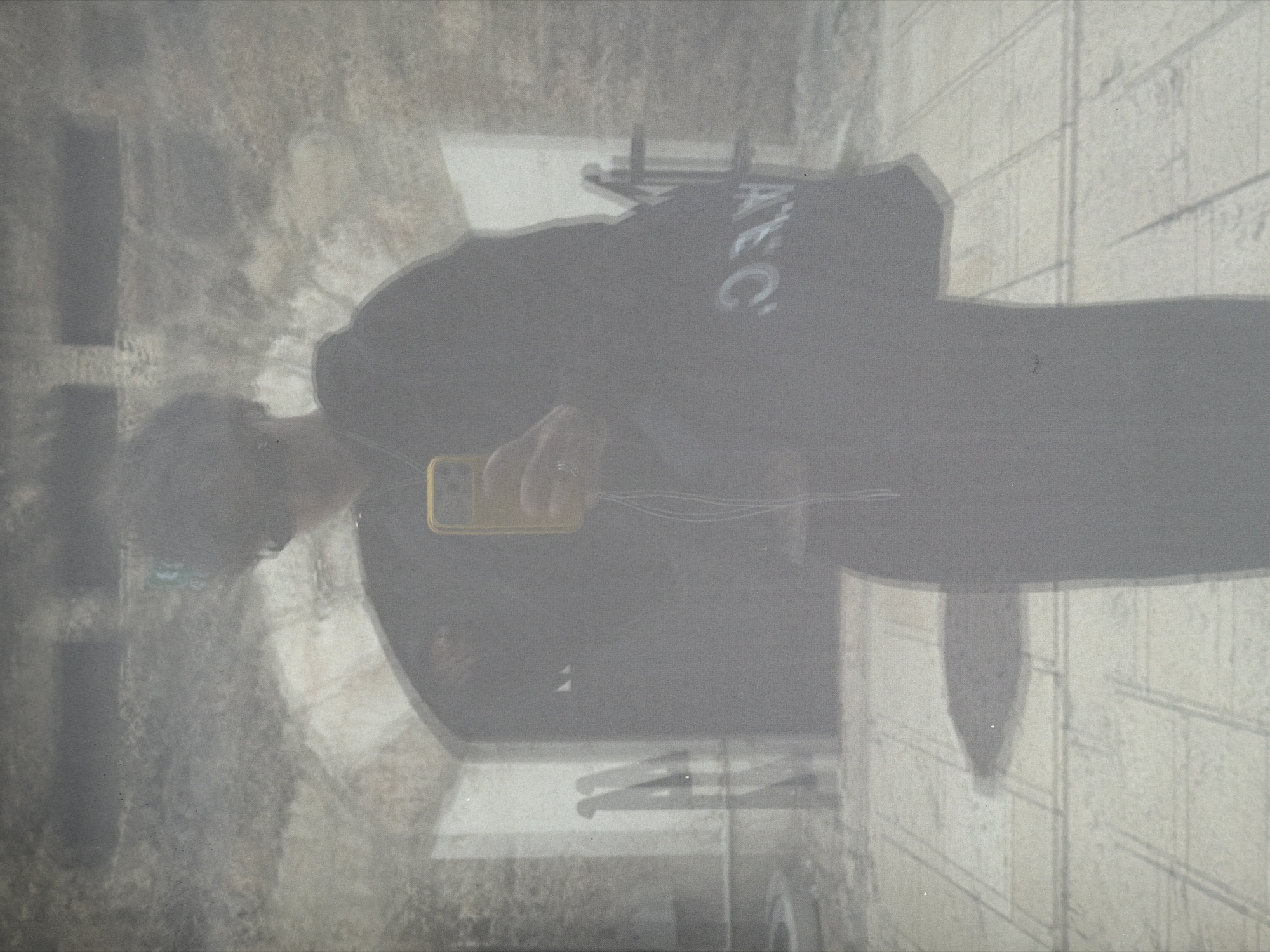 The image shows a person holding a yellow phone, taking a selfie. The person is wearing a dark hooded sweatshirt with "ATEC" written in white on the back. The background is blurred and appears to be outdoors, possibly in an urban setting. The lighting suggests it could be daytime. The image is grainy and has a muted color palette, with shades of gray and beige dominating. The person's face is not clearly visible, but their arm and hand holding the phone are prominent. The overall impression is casual and everyday. There are reflections in the surface, hinting that this might be a photo taken of a reflection in glass or a polished surface. The background has some texture, possibly brickwork or stonework. The image appears to be taken from a low angle, possibly as part of the selfie itself.