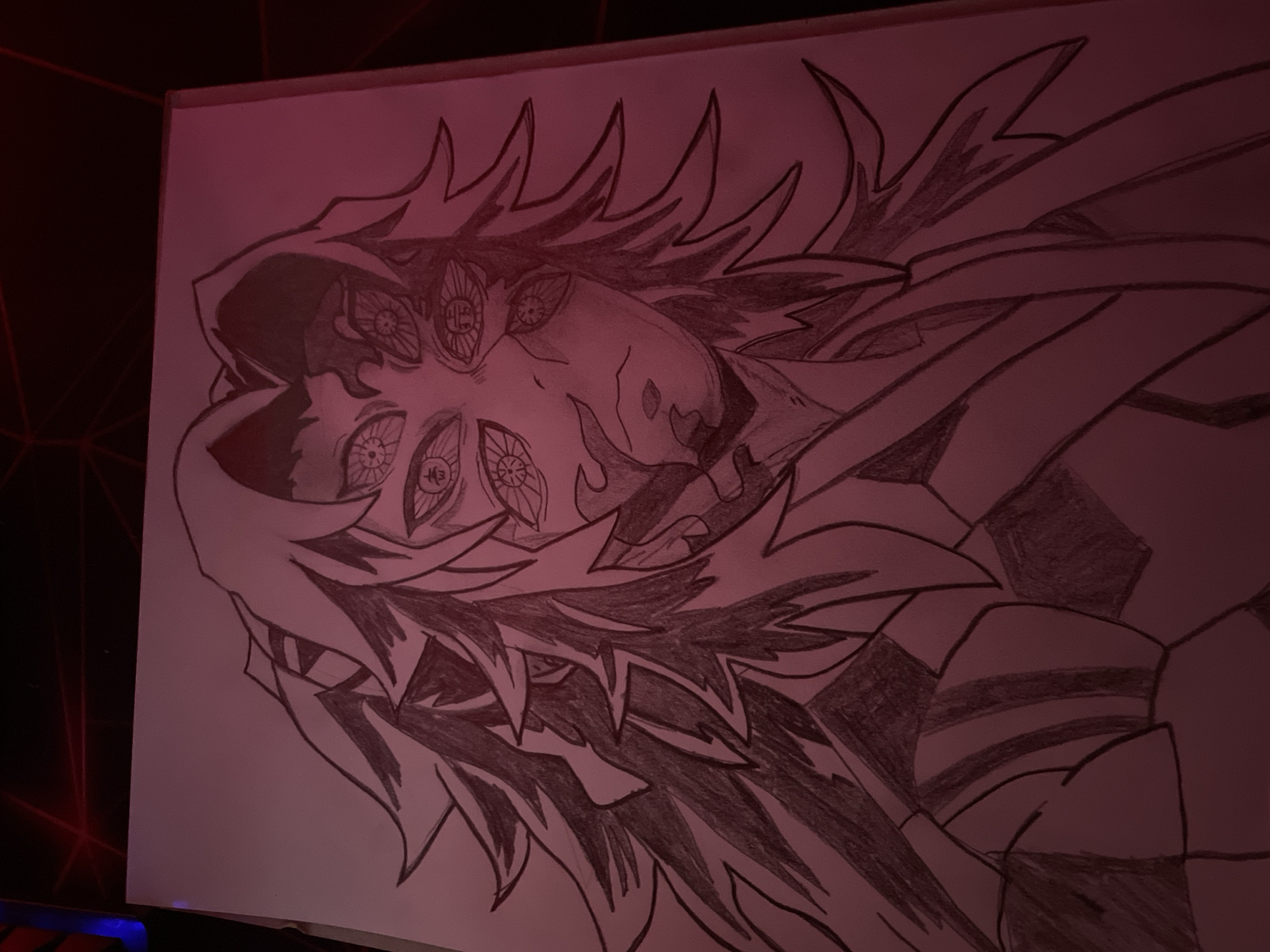 The image depicts a pencil drawing of a character with multiple eyes, sharp features, and spiky hair. The drawing is on a piece of paper, likely a sketchbook or drawing pad. The lighting is dim and appears to have a reddish tint. The character's face is central to the composition. There is no visible information indicating a location in Zlatograd, Bulgaria.