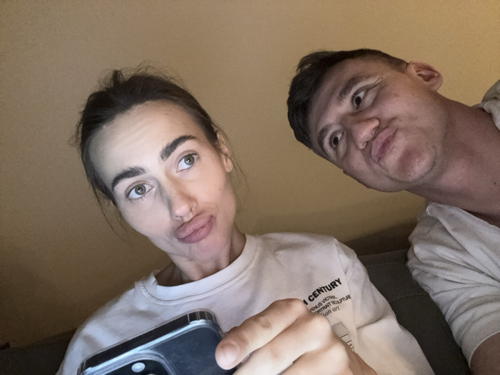 This image depicts two individuals in a casual indoor setting. There are no discernible visual cues within the image to identify its location as Venice, Italy.
A female individual with dark hair, styled in an updo, light-colored eyes, and light skin, occupies the left foreground. She makes a "pouting" facial expression with pursed lips, looking slightly upwards and to her right. She wears a light-colored, long-sleeve top with printed text on the right sleeve, partially visible, which includes "CENTURY", "VENUS VICTRIX", and "SCULPTURE". Her left hand holds a dark smartphone, featuring a visible multi-lens camera array, pointed towards the viewer, indicating a self-portrait (selfie).
Behind and to the right of the female is a male individual with short dark hair and light skin. He also makes a similar "pouting" expression with pursed lips, looking off to his left. He wears a light-colored, short-sleeved top.
The two subjects are posing together for the photograph. The background consists of a plain, light-colored wall or surface. The lighting appears to be dim and localized, possibly emanating from a screen or another close-range light source, casting a soft glow on the subjects' faces.