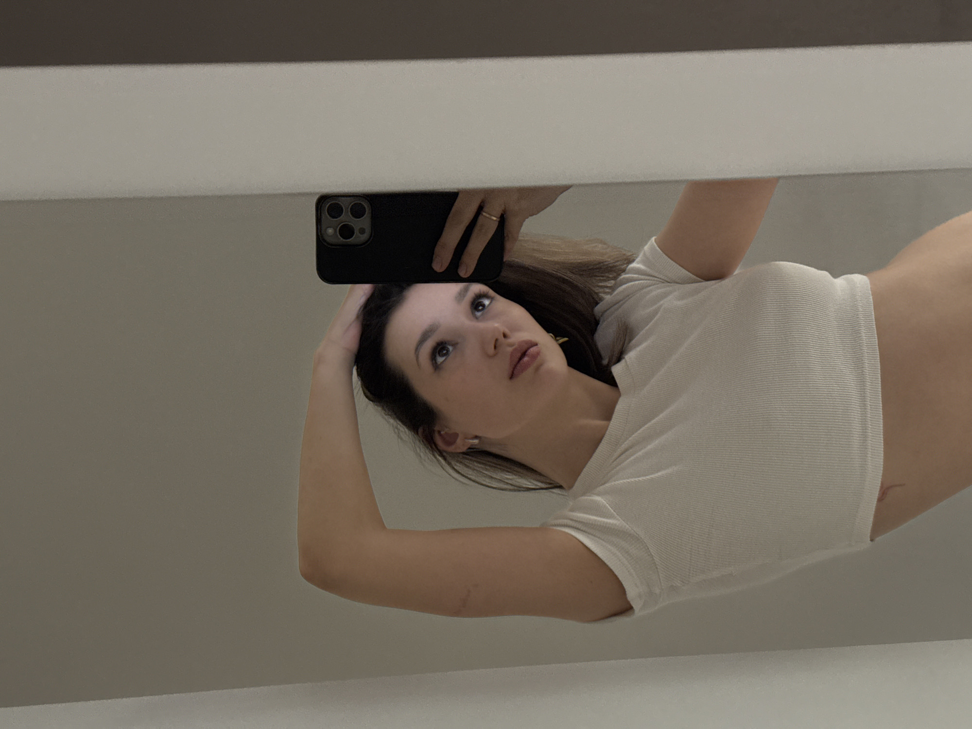 The image displays a young woman taking a selfie reflected in a highly polished, mirror-like surface, possibly a ceiling or large reflective panel. The entire scene is oriented such that the woman appears to be floating horizontally with her head to the left.
The primary subject is a woman with medium-length dark hair, styled down. She is wearing a white, short-sleeved, ribbed crop top, revealing a portion of her midriff on the right side of the frame. Her left arm is bent, with her hand resting gently near her head, while her right hand holds a black-cased smartphone with a prominent triple-lens camera module. A simple band-style ring is visible on her right hand's ring finger. She has an earring in her visible left ear. Her gaze is directed upwards and slightly to the left, towards the phone she's using to capture the reflection. Her expression is calm and contemplative, with subtly defined makeup.
The background, visible around her reflection, is a plain, light grey or beige color, suggesting a minimalist indoor setting. A darker horizontal stripe runs across the top of the image, likely another reflected architectural element. The activity is clearly a self-portrait taken via reflection, utilizing the unique angle and surface. There is no visible text in the image.