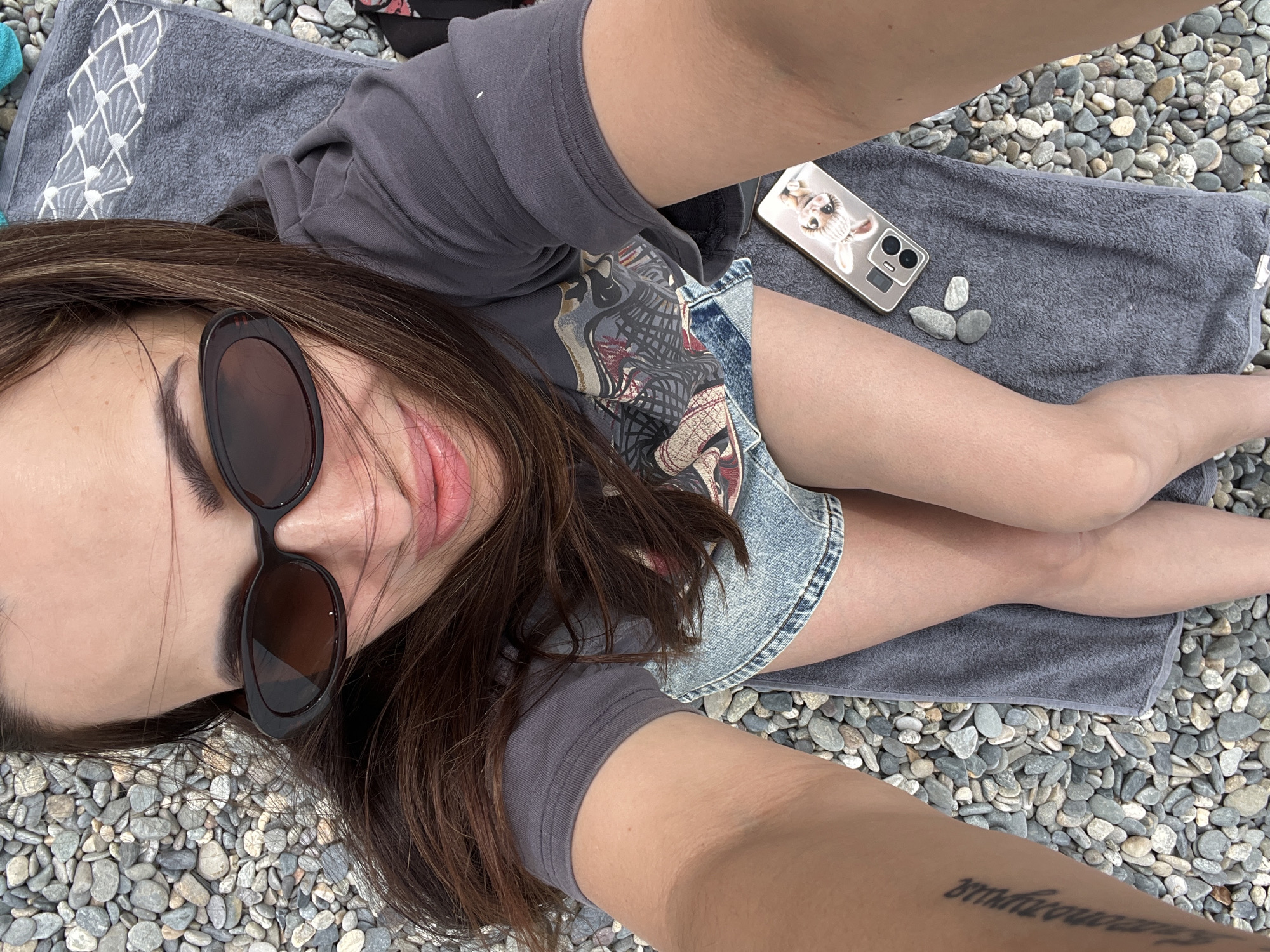 A top-down selfie shows a young woman lying on a grey towel on a pebbly beach. She is wearing sunglasses and a dark t-shirt over a denim skirt. Her legs are crossed, and her phone, with a cartoon rabbit case, is placed on the towel beside her. The background is filled with grey and white pebbles. The lighting suggests it is daytime.