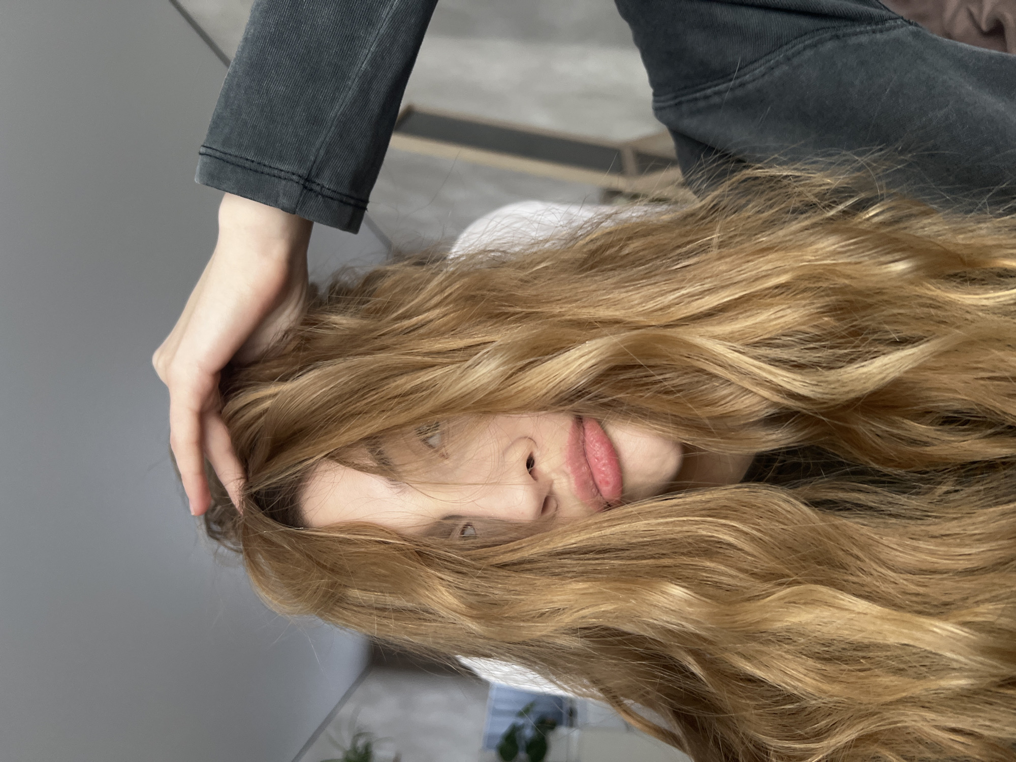 A woman with long, wavy, blonde hair is positioned in the center of the frame, her face visible amidst the strands. She is wearing a white shirt and grey pants, and her left hand is touching her hair. The background consists of a grey wall and a glimpse of an interior space with a plant. The location is Unknown, Unknown.