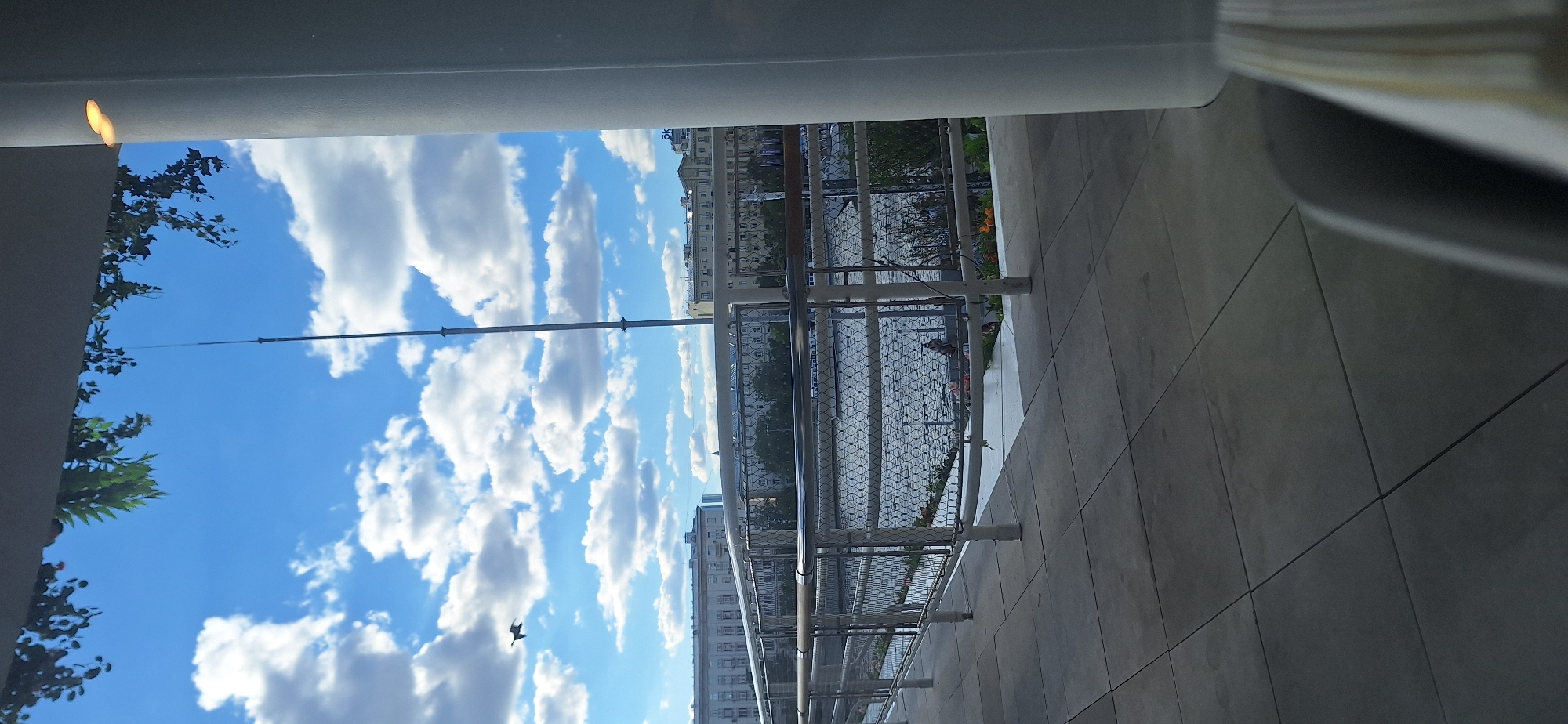 The image, taken from a high vantage point, possibly a balcony or upper floor of a building, shows a view of Moscow, Russia on a sunny day with fluffy white clouds in a blue sky. The camera is pointed toward an area of the city featuring other buildings, trees, and an unidentified body of water. A metal fence or railing is visible in the foreground, and a bird is visible in the sky. The floor of the balcony or vantage point is composed of large square tiles.