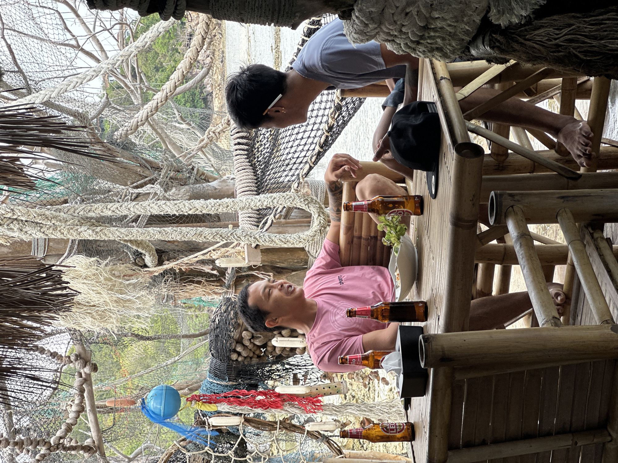 Two men are seated at a bamboo table outdoors, with a body of water visible in the background. The man on the left, wearing a pink t-shirt, is looking to his right and appears to be speaking. He has tattoos on his arm. The man on the right, wearing a grey t-shirt, is looking to his left and has a cigarette behind his ear. A black baseball cap rests on the table in front of him. On the table, there are three bottles of beer, a tissue box, a bowl of food, and some green leaves. The surroundings are rustic and decorated with fishing nets, ropes, and buoys, suggesting a coastal or beachfront establishment. The overall scene depicts a casual outdoor dining or drinking setting in ตำบลเกาะพะงัน, Thailand.