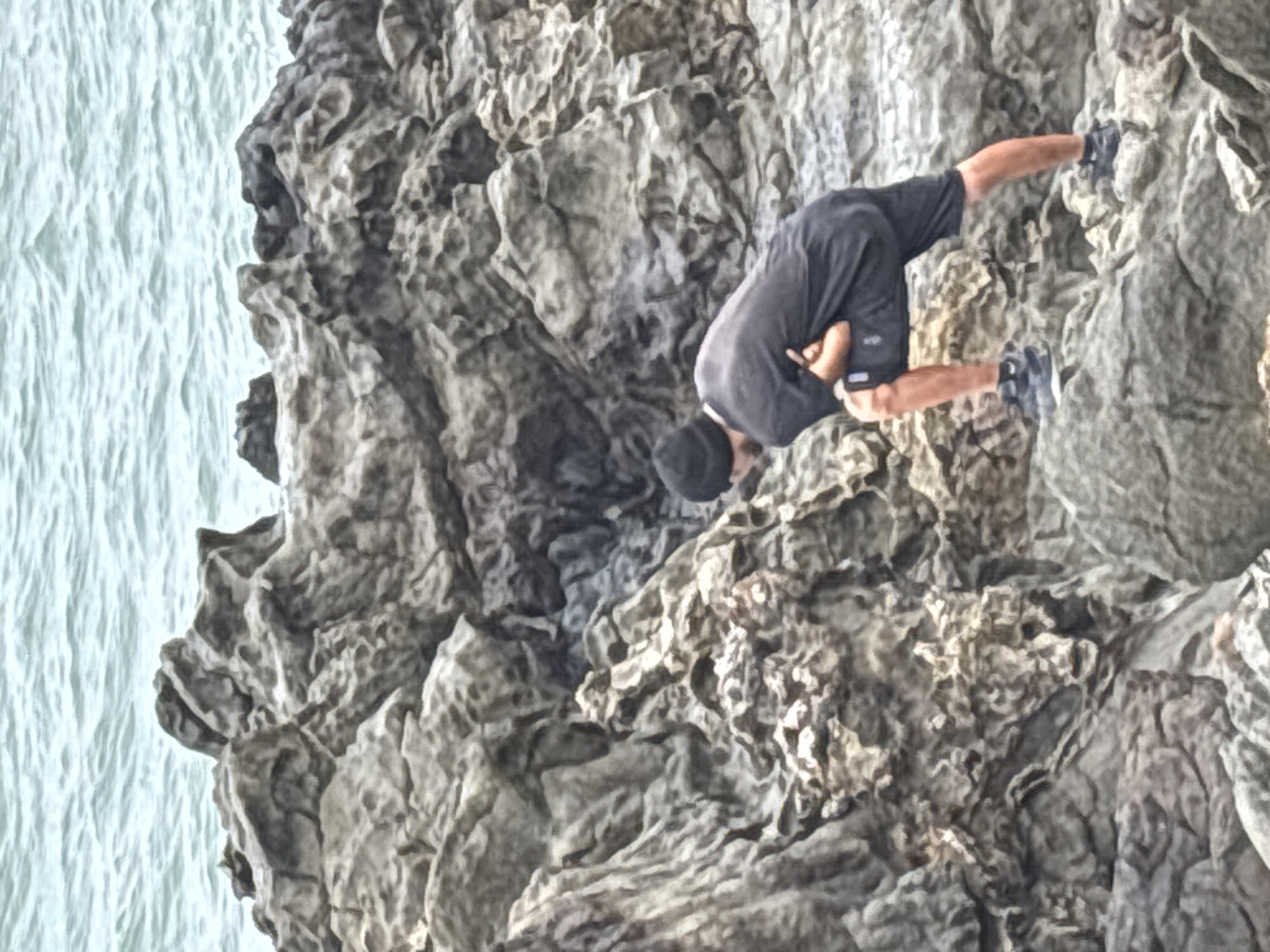 A man wearing dark clothing is rock climbing on a cliff face. He is wearing a dark t-shirt, dark shorts, a dark hat, and climbing shoes. He is positioned on the rock with his legs higher than his head. The cliff is grey and textured. To the left of the cliff face is a body of water. This is likely in Arica, Chile, given the location information provided.