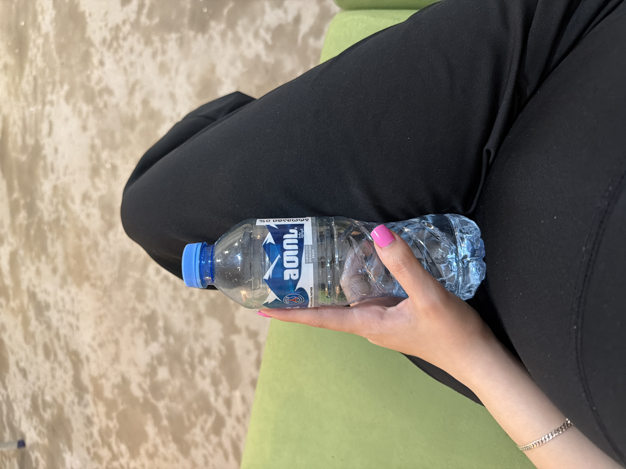 A person's arm and hand hold a clear plastic bottle of water. The bottle has a blue cap and a label with blue and white mountain imagery, a red "Paris" logo, and text in Georgian. The person is wearing black pants and a silver bracelet. The background shows a green upholstered surface and a textured, light-colored carpet. The scene appears to be indoors.