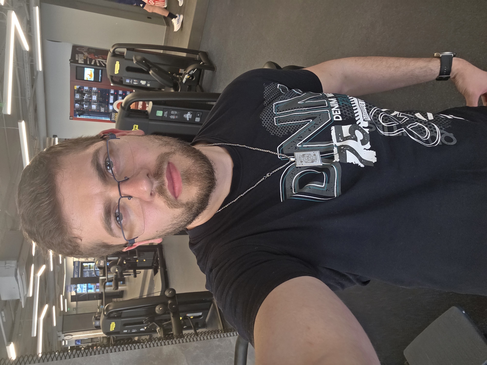A selfie, taken indoors, likely in a gym. A Caucasian male with a beard and glasses, wearing a black T-shirt with a print, a silver chain with a pendant, and a wristwatch, is the primary subject. Background shows gym equipment (including fitness machines and a vending machine). Location unidentifiable as Plovdiv, Bulgaria based on the image.