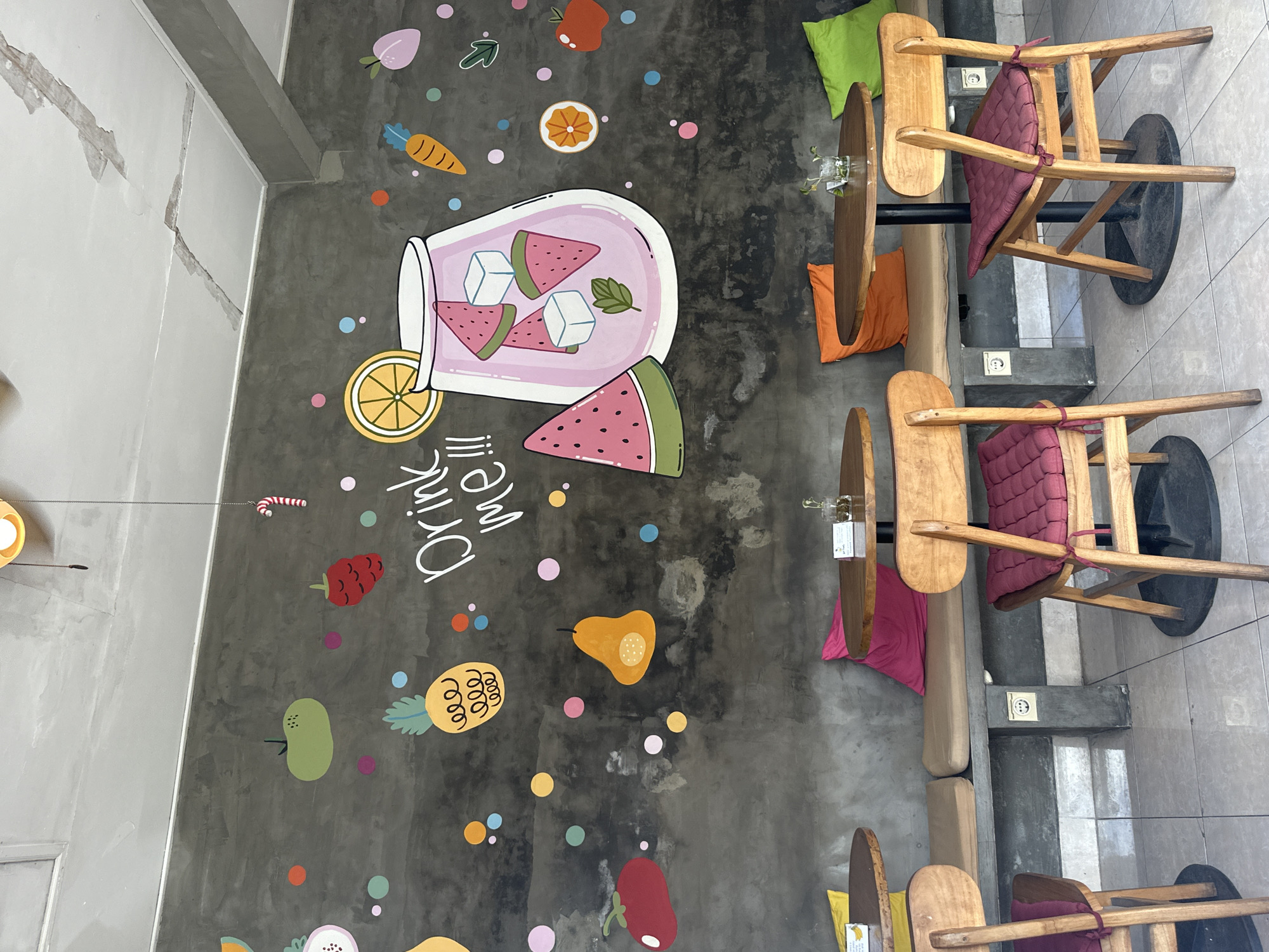 The image shows an interior shot of a cafe in Ubud District, Indonesia, featuring a dark gray concrete wall adorned with colorful fruit-themed artwork and playful dots. Dominating the wall are illustrations of a large glass of pink juice filled with watermelon slices and ice cubes, a sliced watermelon, a pineapple, a tomato, a pear, a carrot, a strawberry, and a slice of orange. Various colorful dots are scattered around the artwork, adding to the whimsical atmosphere.
In the foreground, two round wooden tables with black bases are visible, each accompanied by two wooden chairs with maroon cushions. One table is positioned in the center-left of the frame, and another is in the center-right. A long, built-in bench runs along the base of the wall, with brown cushions. Brightly colored pillows in pink, orange, and green are placed on the bench.
The flooring consists of light-colored square tiles. The ceiling is white with visible cracks and repairs. A light fixture with a yellow shade hangs from the ceiling. The overall impression is a vibrant and casual dining or resting space. No people are present in the image.