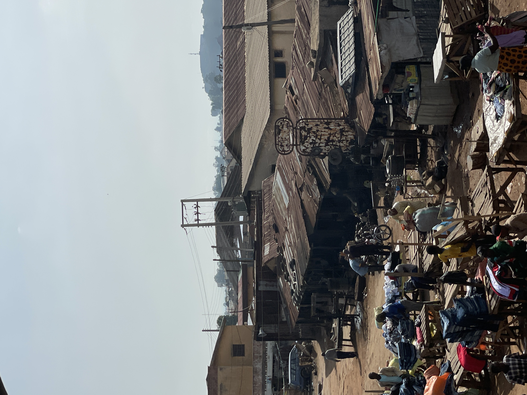 The image shows an outdoor market in Bukuru, Nigeria. There are multiple people browsing and selling merchandise, primarily clothing. The scene is densely packed with wooden stalls and displays. The architecture includes buildings with corrugated metal roofs, some rusted. Power lines are visible overhead. In the background, there is a hill with a structure on top. The overall scene is dusty and suggests a crowded, bustling commercial area.