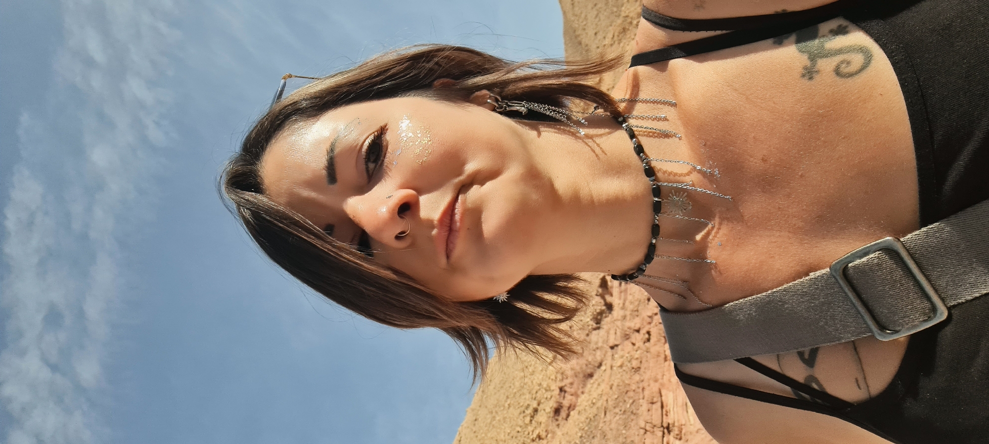 A woman is standing outside, with the sky in the background. She has short brown hair and is wearing a black top, a black beaded necklace with silver charms, and a grey strap over her shoulder. She has a nose ring and shimmering makeup on her face. A tattoo of a lizard is visible on her upper chest. The location is likely near a desert area, as indicated by the sandy-colored ground in the background. The city is Hevel Eilot Regional Council, Israel.