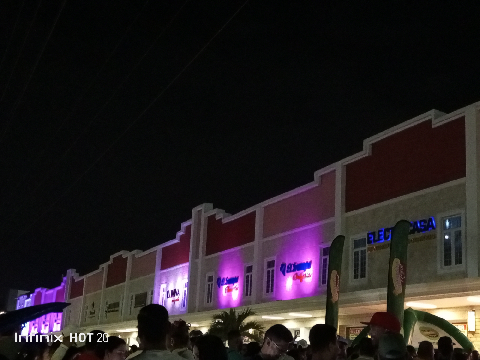 At night in Porlamar, Venezuela, a crowd of people stands in front of a row of businesses. The buildings are two-storied, with reddish-brown tops and beige facades, featuring large, bright signs in neon colors of purple and blue. Palm trees are interspersed among the crowd. Inflatable promotional items are visible. Power lines run across the top of the image against a dark sky. The scene appears to be at an outdoor gathering or event.