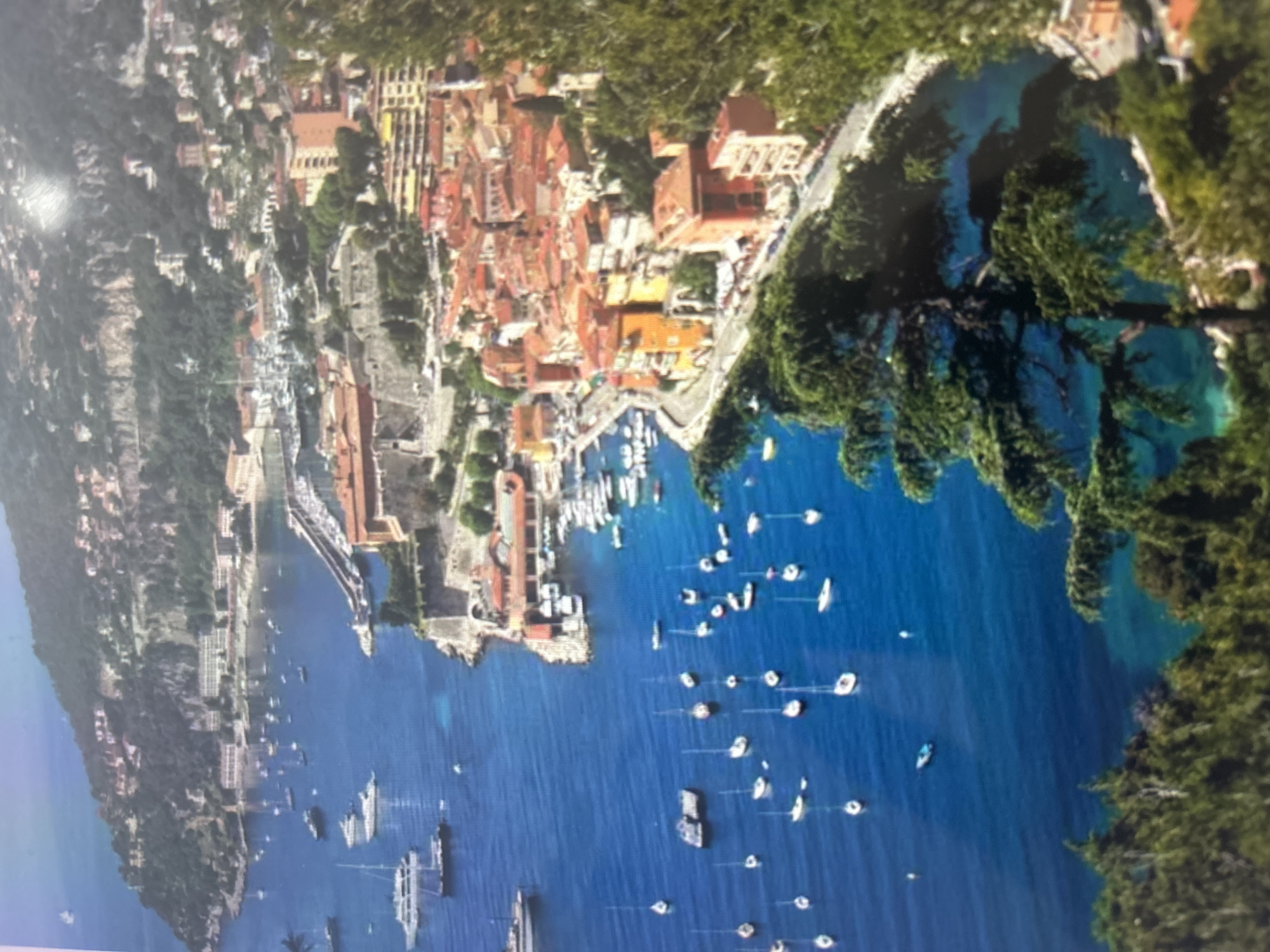An aerial, high-angle view captures a picturesque coastal town nestled along a deep blue bay. The image is set on a clear, sunny day, likely during late morning or early afternoon, with vibrant illumination.
The main subject is a densely built town with numerous buildings featuring distinctive terracotta or reddish-orange tiled roofs. This architecture is characteristic of the Mediterranean region. The town cascades down a hillside towards the water, with a prominent harbor or marina in the foreground, bustling with various boats. These include many white recreational yachts, sailboats, and motorboats anchored or docked in the clear, deep blue waters of the bay. Some larger vessels are visible further out in the bay.
Lush green vegetation, including dense trees and foliage, covers the surrounding hills and mountains, providing a vibrant contrast to the urban structures and the blue sea. The coastline features rugged terrain with cliffs and wooded areas, framing the tranquil waters.
Towards the left side of the image (as if oriented correctly), a long stretch of coastline continues, showing more buildings and a distinct bridge or viaduct structure extending across a portion of the landscape, suggesting significant infrastructure. The water in the bay is exceptionally clear, with lighter turquoise hues visible closer to the shore in some areas.
The scene depicts a peaceful and beautiful coastal settlement, possibly a popular resort or residential area, with no specific human activity clearly discernible due to the distance. The overall impression is one of serene beauty and Mediterranean charm. No visible text is present in the image. This location strongly resembles Villefranche-sur-Mer on the French Riviera.