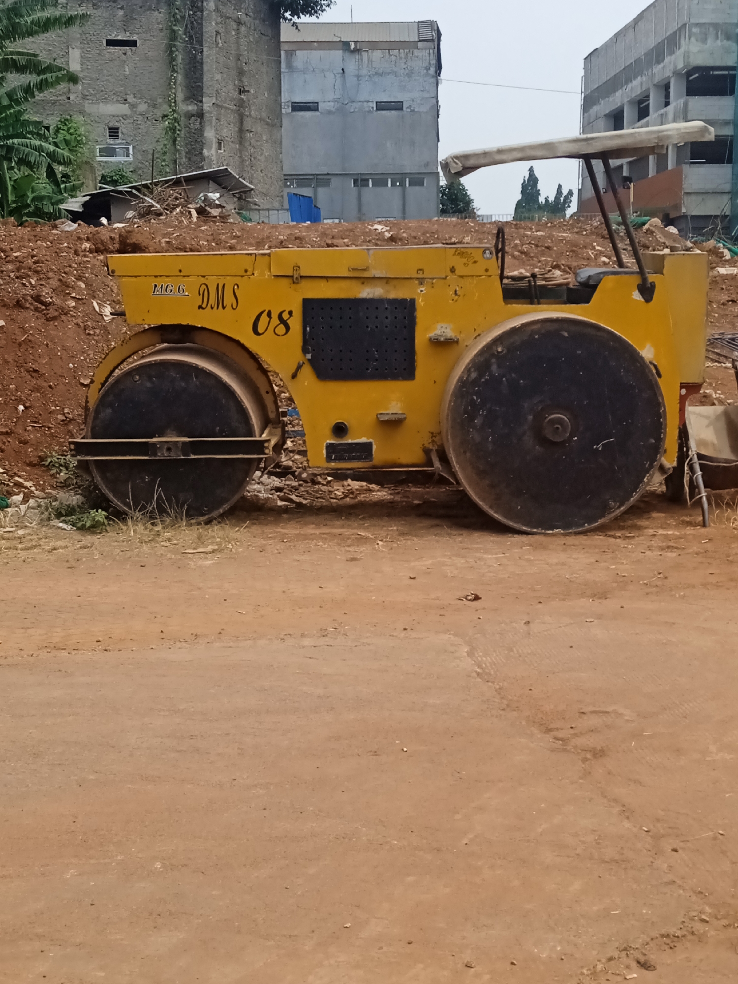 A yellow steamroller sits on a dirt construction site, likely in Bekasi, Indonesia. The steamroller is positioned horizontally in the center of the frame, with its large metal roller in the foreground. The roller is dark gray and appears to be made of solid metal. Above the roller, the yellow body of the steamroller is visible, with the text "M.G.C. DMS 08" printed in black on its side. The machine has a canvas canopy supported by metal poles, suggesting it is an older model.
Behind the steamroller, the landscape is uneven and covered in brown dirt and debris. In the background, there are several buildings under construction. One building on the left is made of weathered gray stone with visible vines growing on its facade. Another building to the right is a modern concrete structure with multiple floors and exposed windows. The sky is overcast and a pale gray. There are no people visible in the image. The overall impression is one of an active construction site in an urban or semi-urban area.