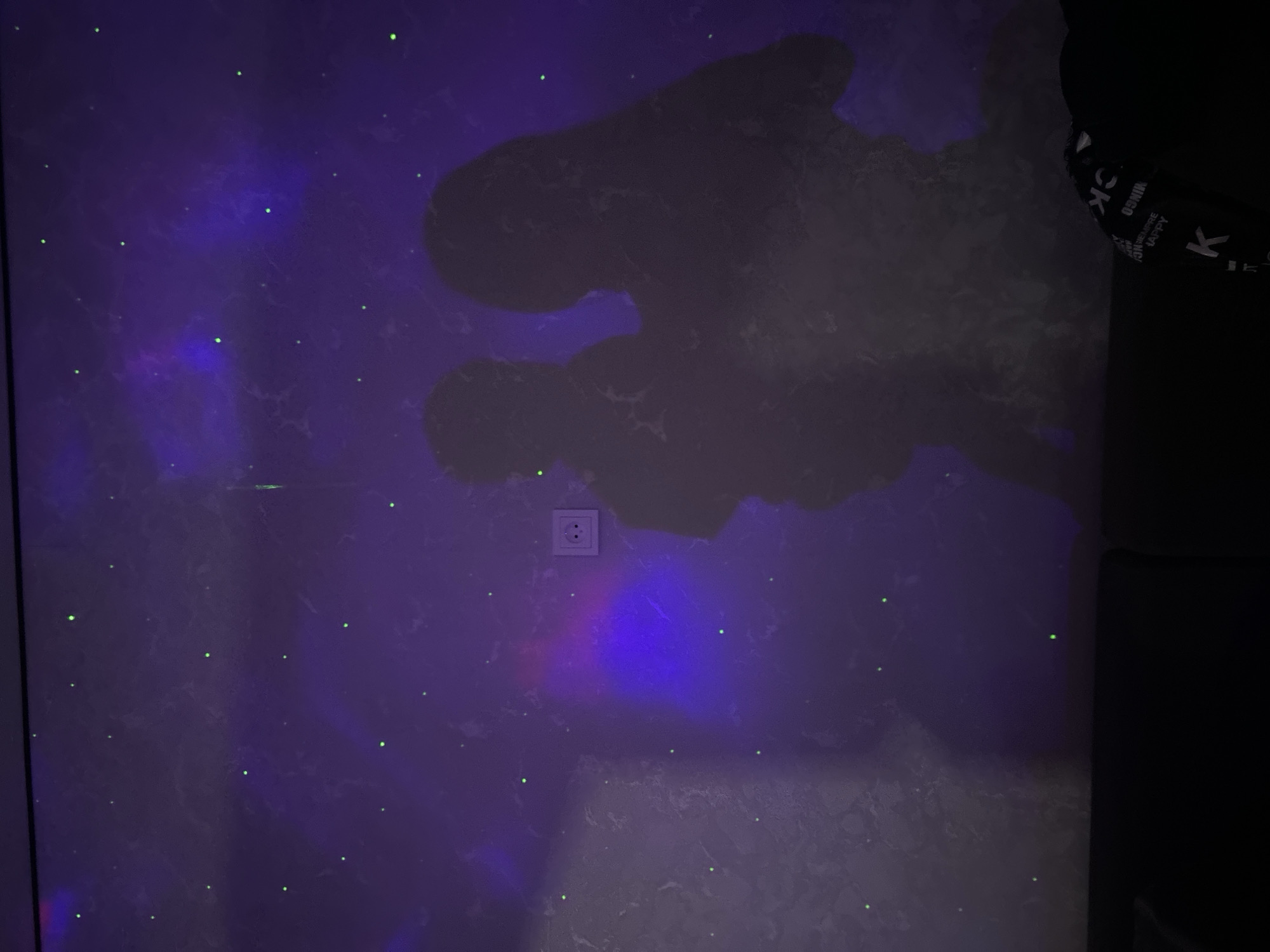 The image shows a wall with a gray marble-like pattern. There are numerous small green dots scattered across the wall, resembling stars. A large, dark, amorphous shadow covers a significant portion of the upper right area. An electrical outlet is visible on the wall. A dark object intrudes from the right edge of the frame, obscuring the wall. Based on previous context, this image is located in Chaikovsky, Russia.
